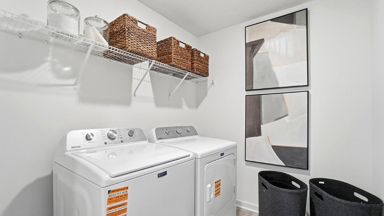 Laundry room with white top-loading washer and dryer, wire shelf with woven baskets, wall art, and two hampers.