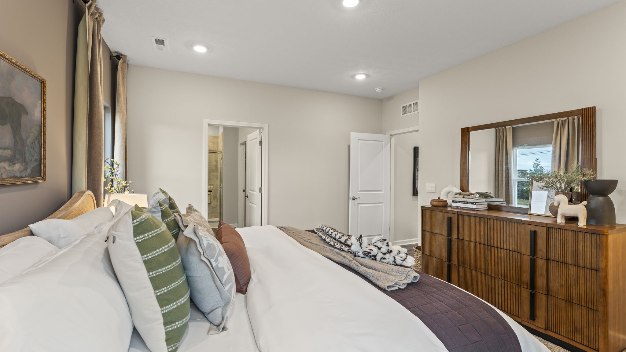 Closeup of primary bed with primary bathroom and wooden dresser in view.