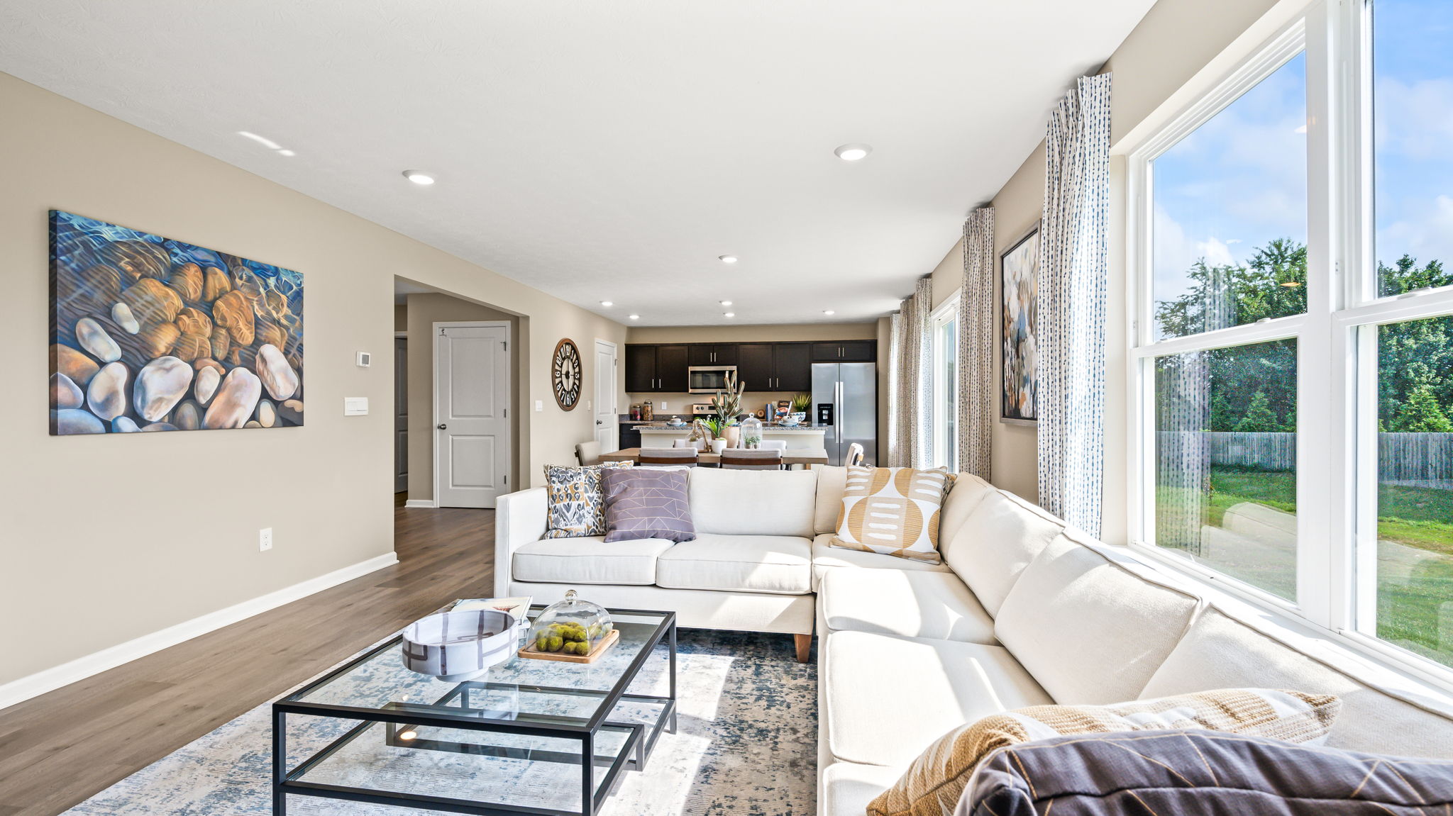 Sunlit living room with a white sectional, glass coffee table, large windows, and views toward the dining area and kitchen.
