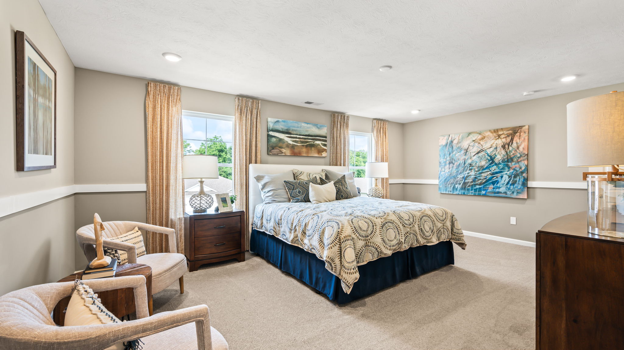 Large bedroom with a patterned bed, dark wood dressers and nightstands, paired chairs, multiple windows, and framed artwork.