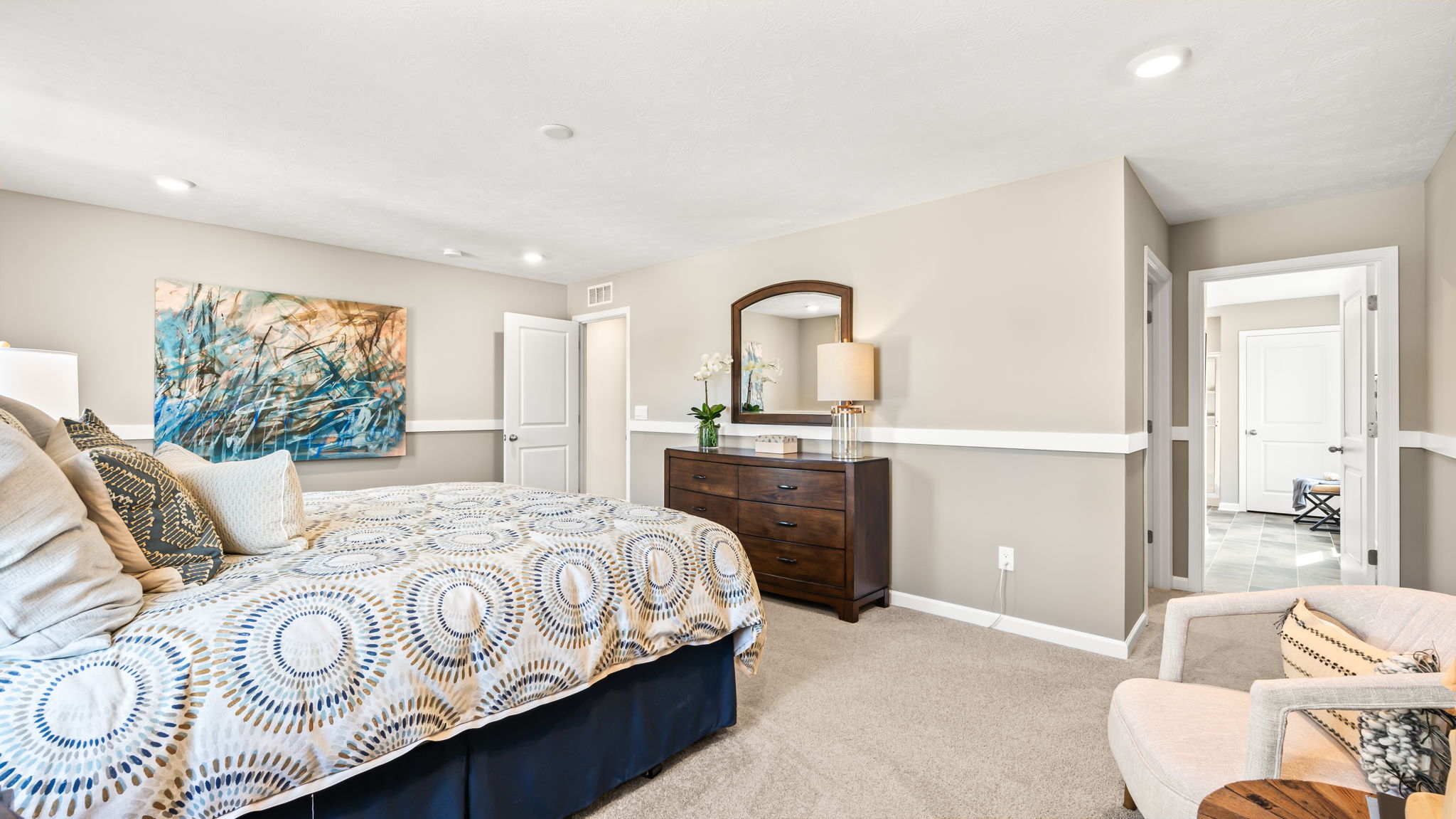 Bedroom view across a patterned bed toward a dark wood dresser, arched mirror, abstract artwork, chair, and bathroom doorway.