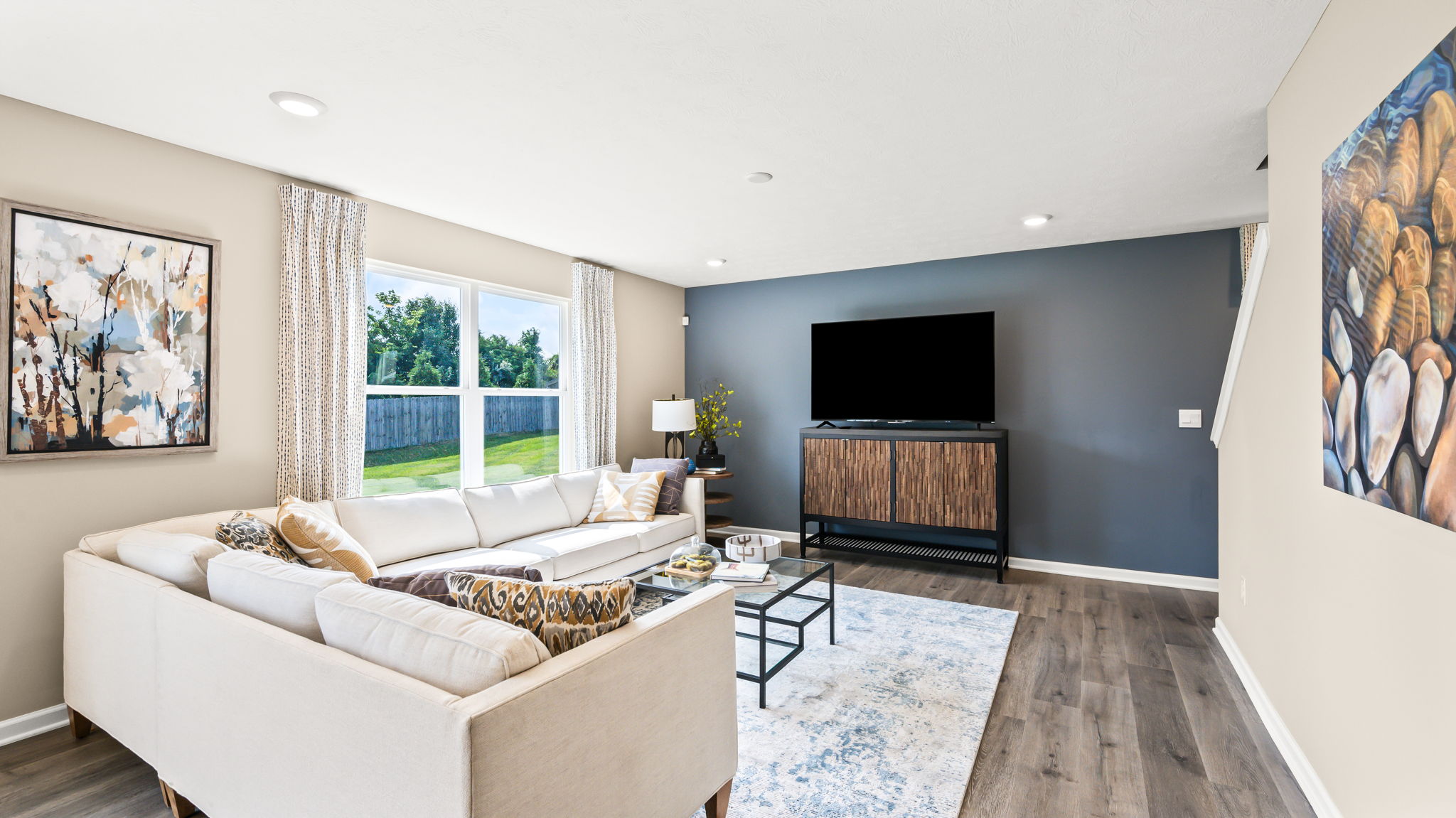 Living room with a white sectional, glass coffee table, large windows, and a wall-mounted television on a dark blue accent wall.