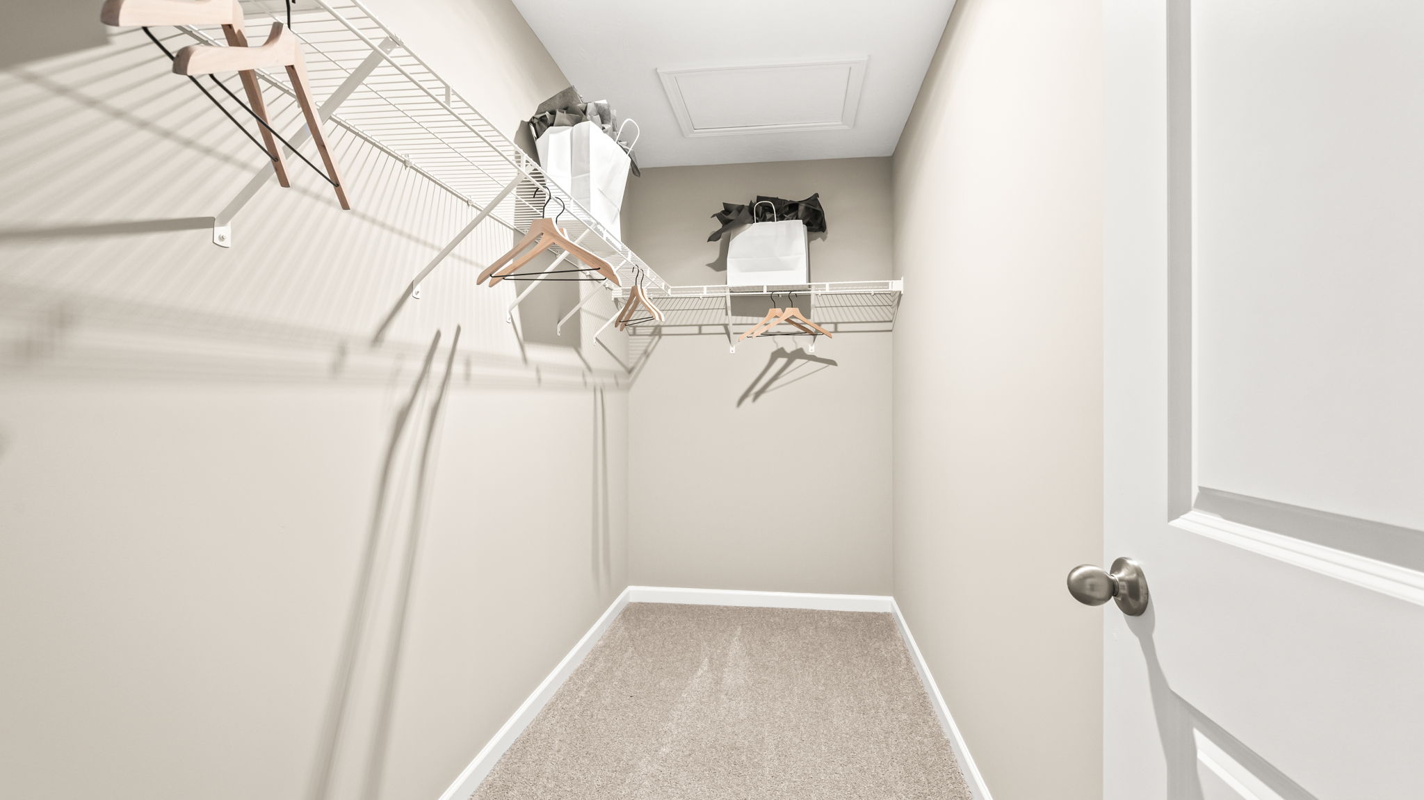 Walk-in closet with beige walls and carpet, white wire shelving, wooden hangers, storage bags, and an open paneled door.