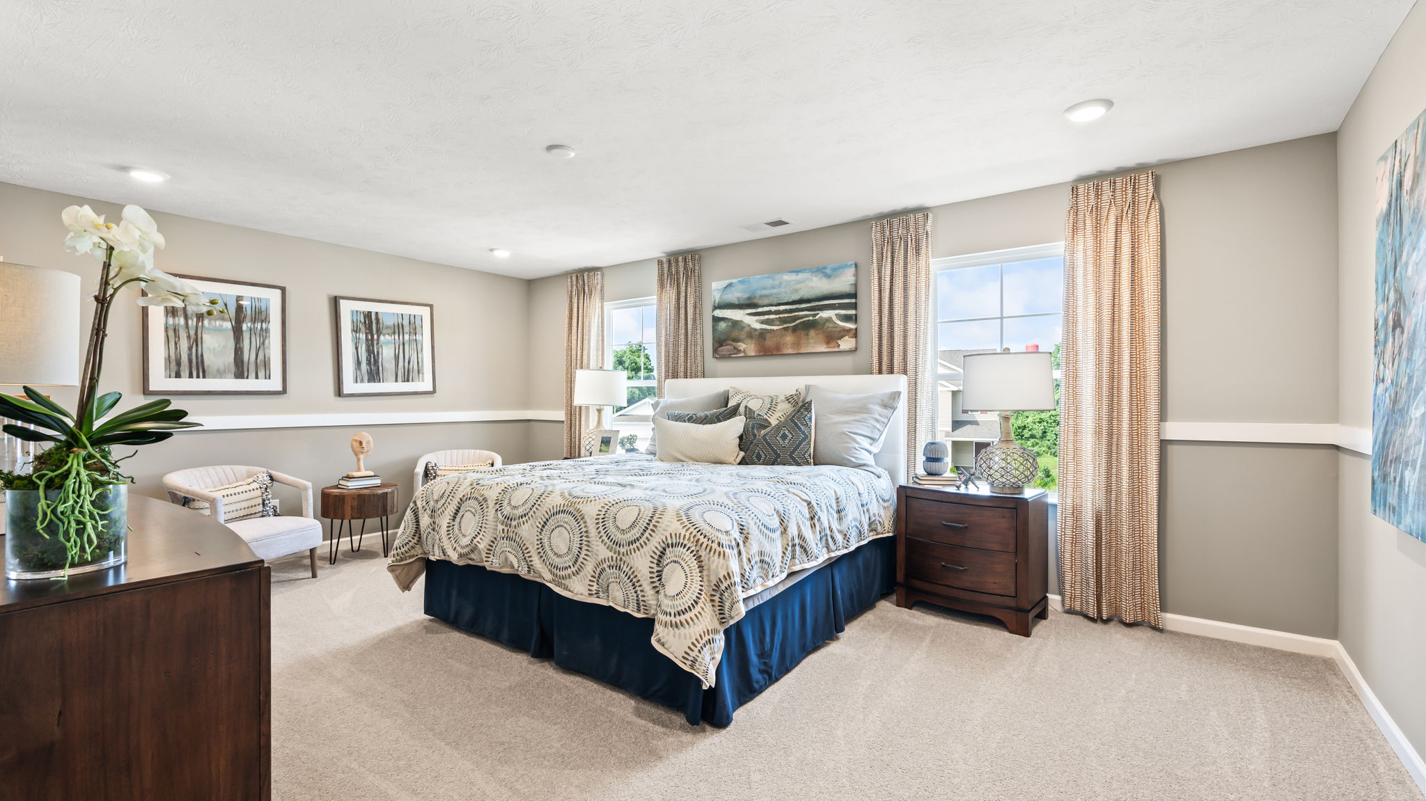 Spacious bedroom with a patterned bed, dark wood nightstands, two windows with curtains, framed art, and a seating area.