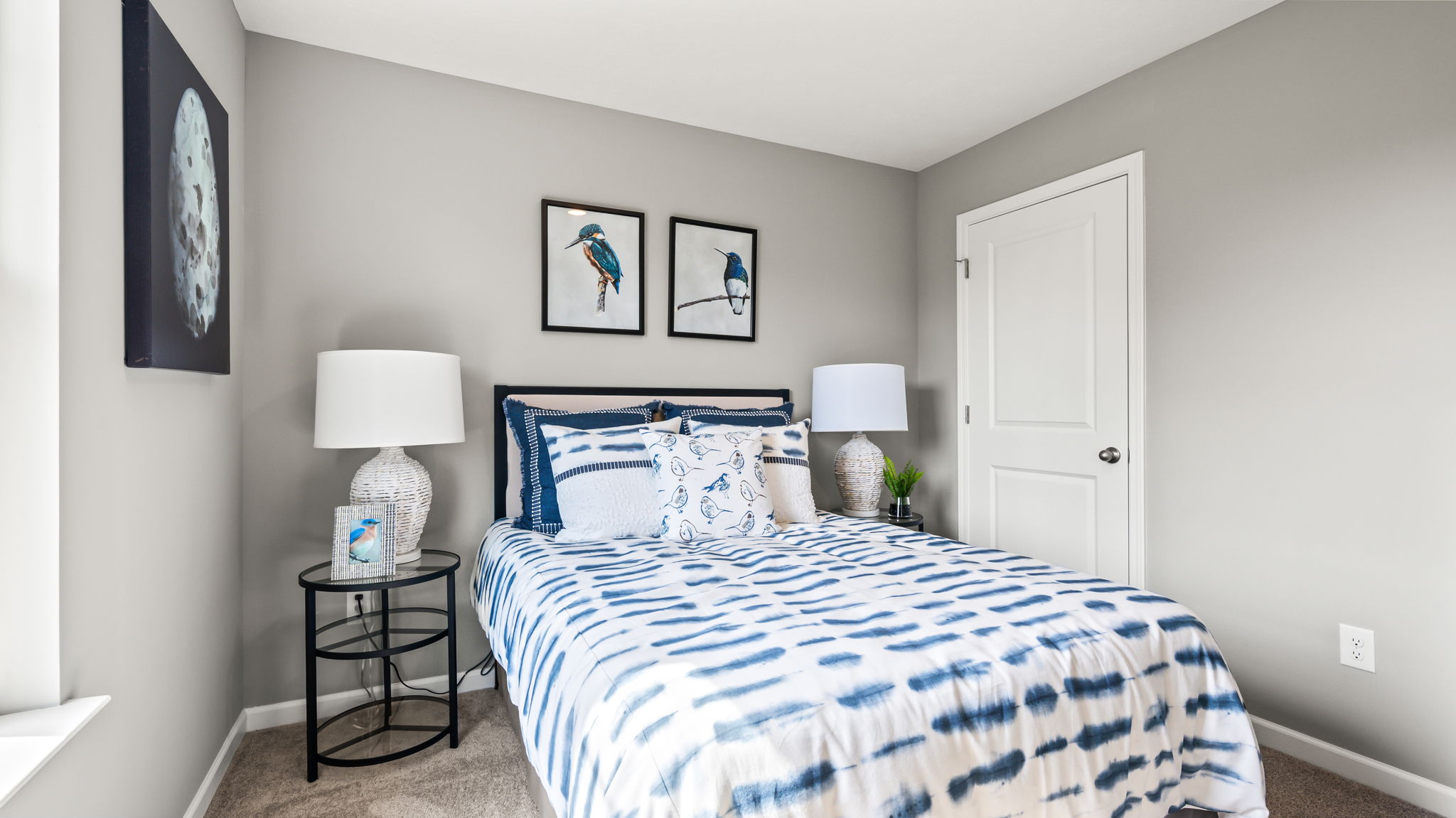 Gray bedroom with blue-and-white patterned bedding, bird and egg artwork, dark wood dresser, window, and white lamps.
