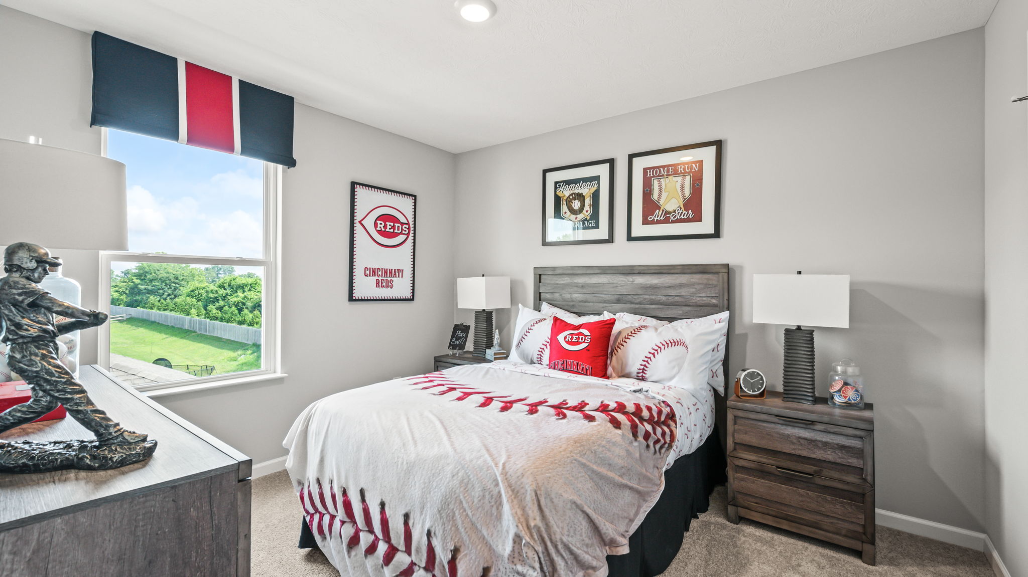 Bedroom view across blue-and-white bedding toward a wood dresser, round black mirror, white lamp, plant, and open hallway door.