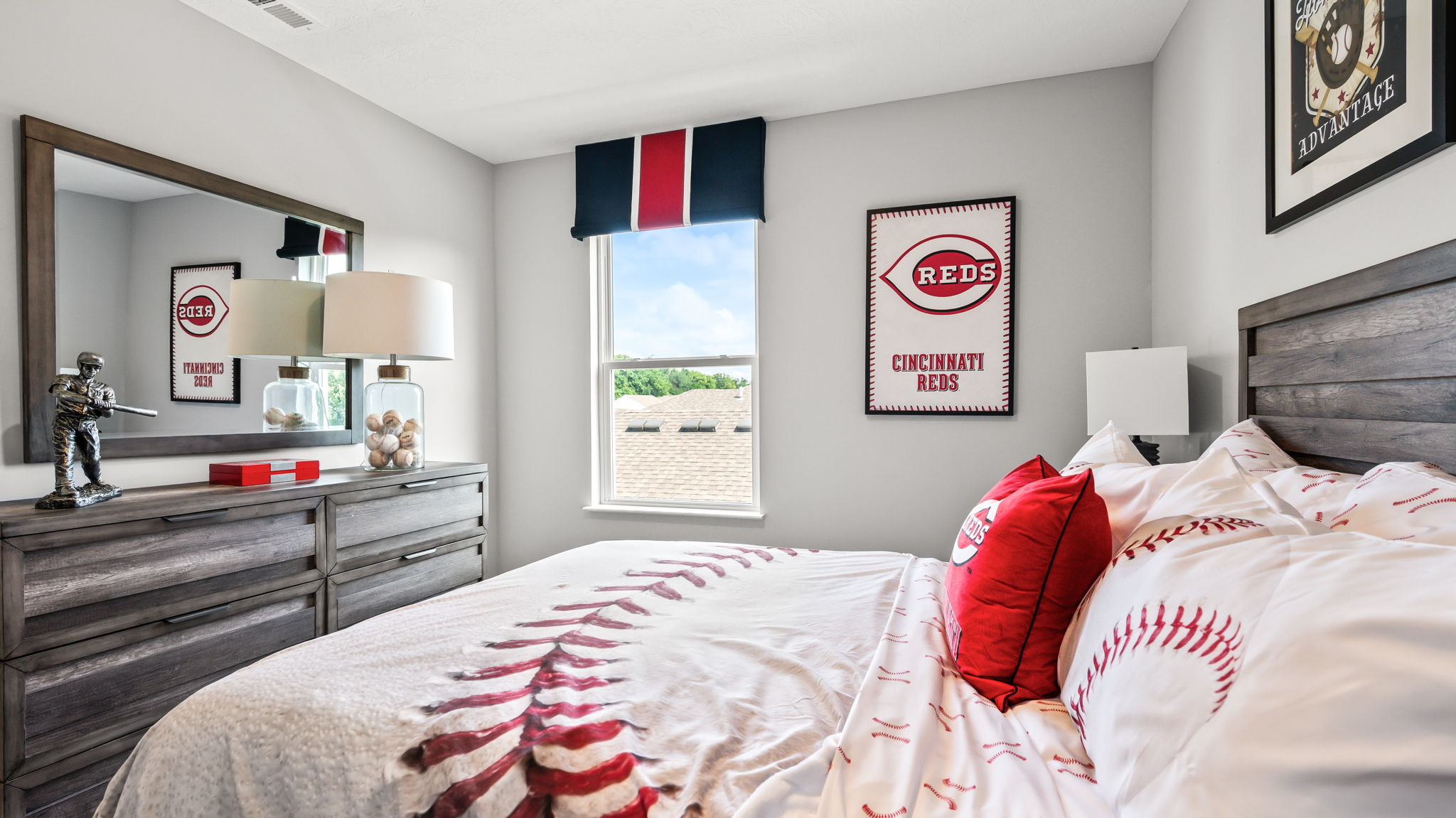 Baseball-themed bedroom with Cincinnati Reds bedding and wall art, gray wood furniture, matching lamps, and a window overlooking the yard.