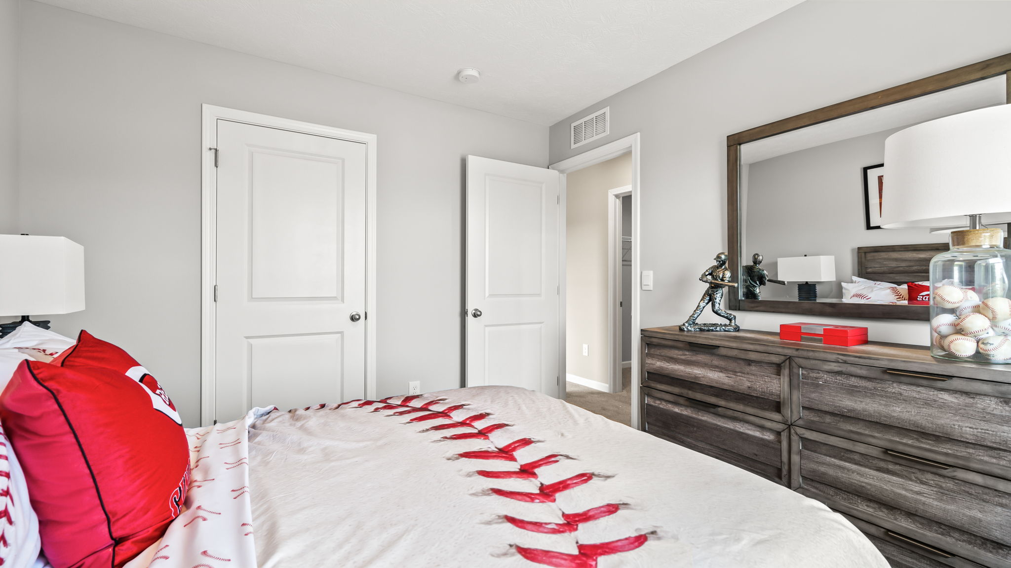 View across a baseball-patterned bed toward a gray wood dresser and mirror decorated with a batter figurine, baseballs, and red accents.