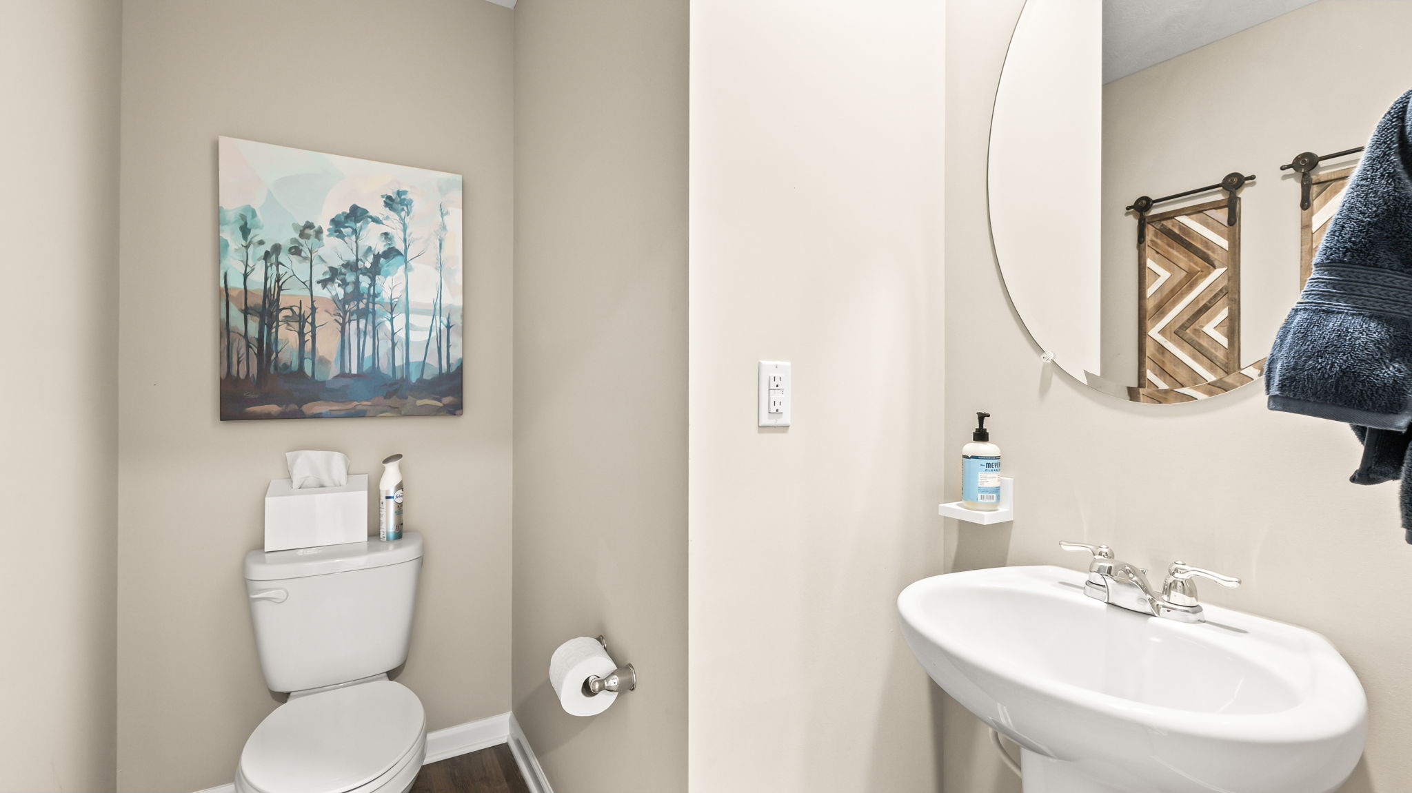 Powder room with a white pedestal sink, round mirror, toilet, towel rack, neutral walls, and blue forest artwork.