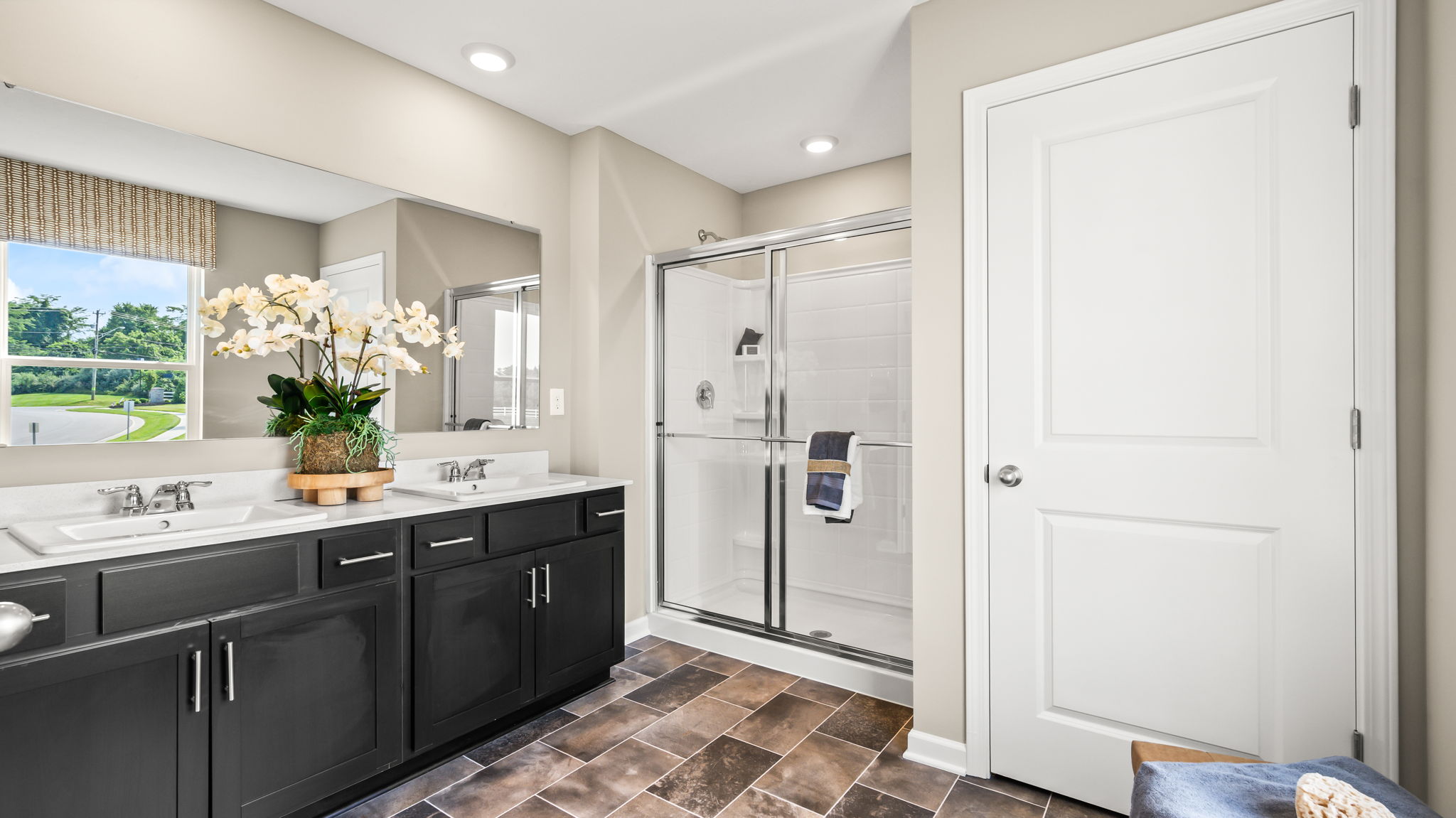 Bathroom with a dark double-sink vanity, large wall mirror, glass-enclosed shower, tiled floor, and white paneled door.