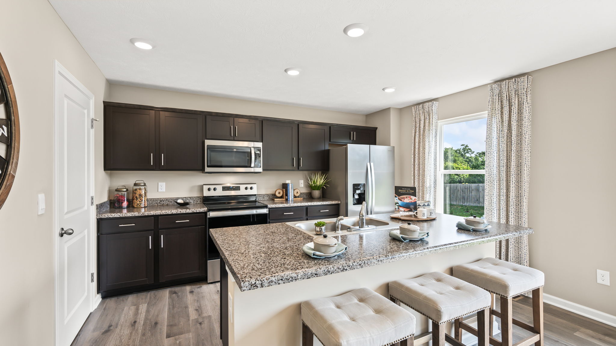 Wide kitchen view with dark cabinets, stainless steel appliances, granite island with double sink, and three upholstered stools.
