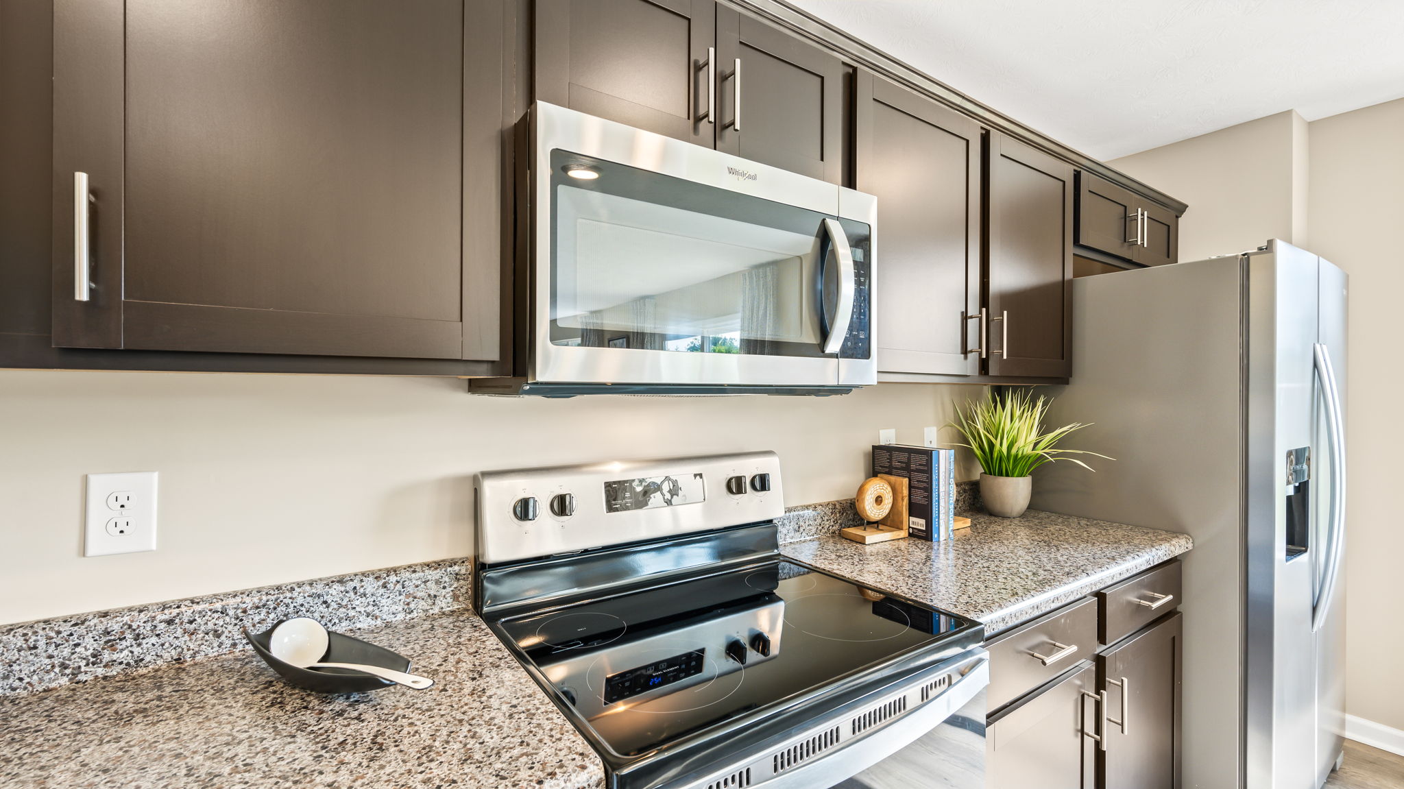 Close view of the kitchen range and microwave set between dark cabinets, with granite counters, books, a plant, and refrigerator.