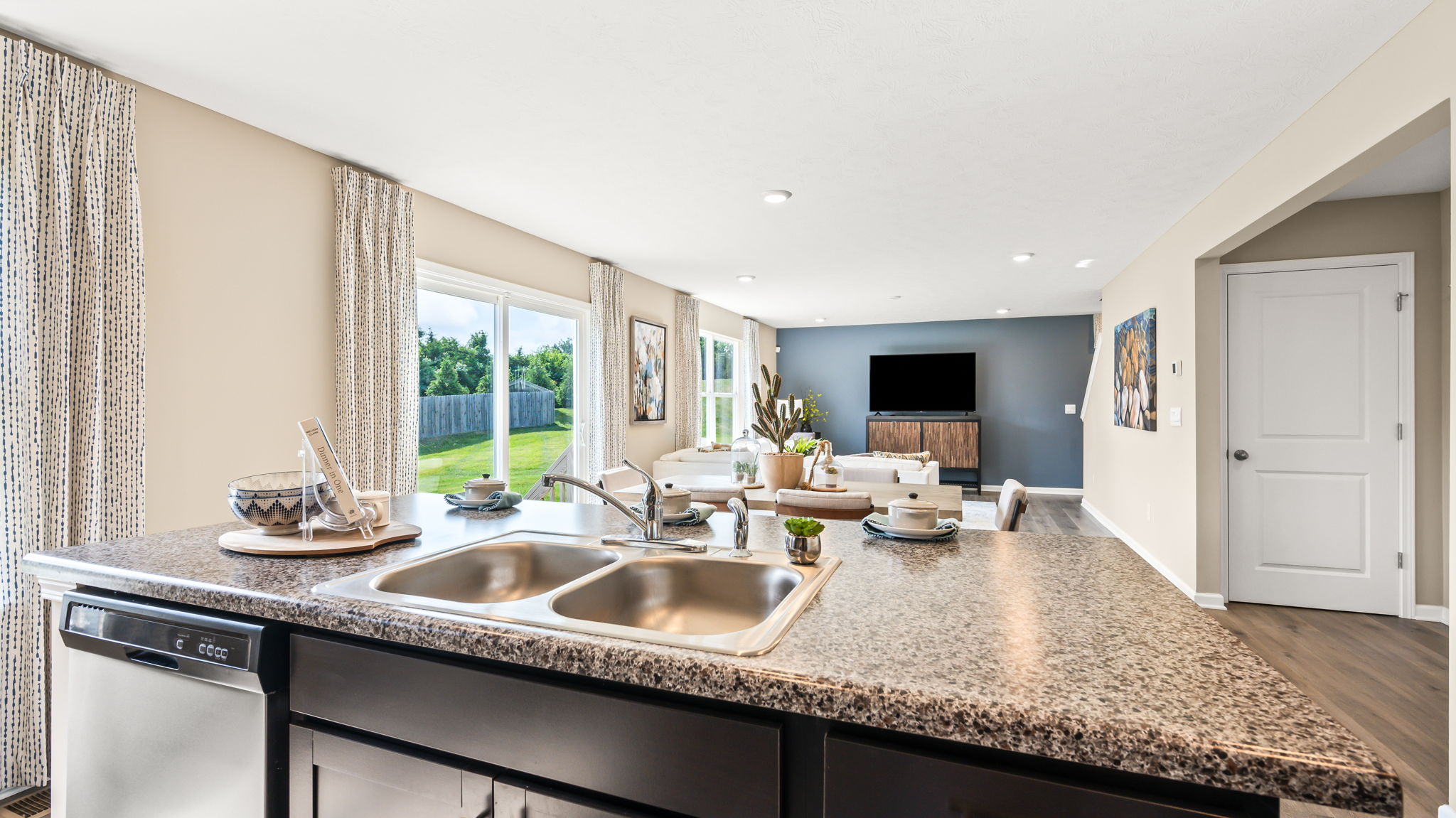 View across the kitchen island with double sink toward the open living room, large windows, white seating, and a television wall.