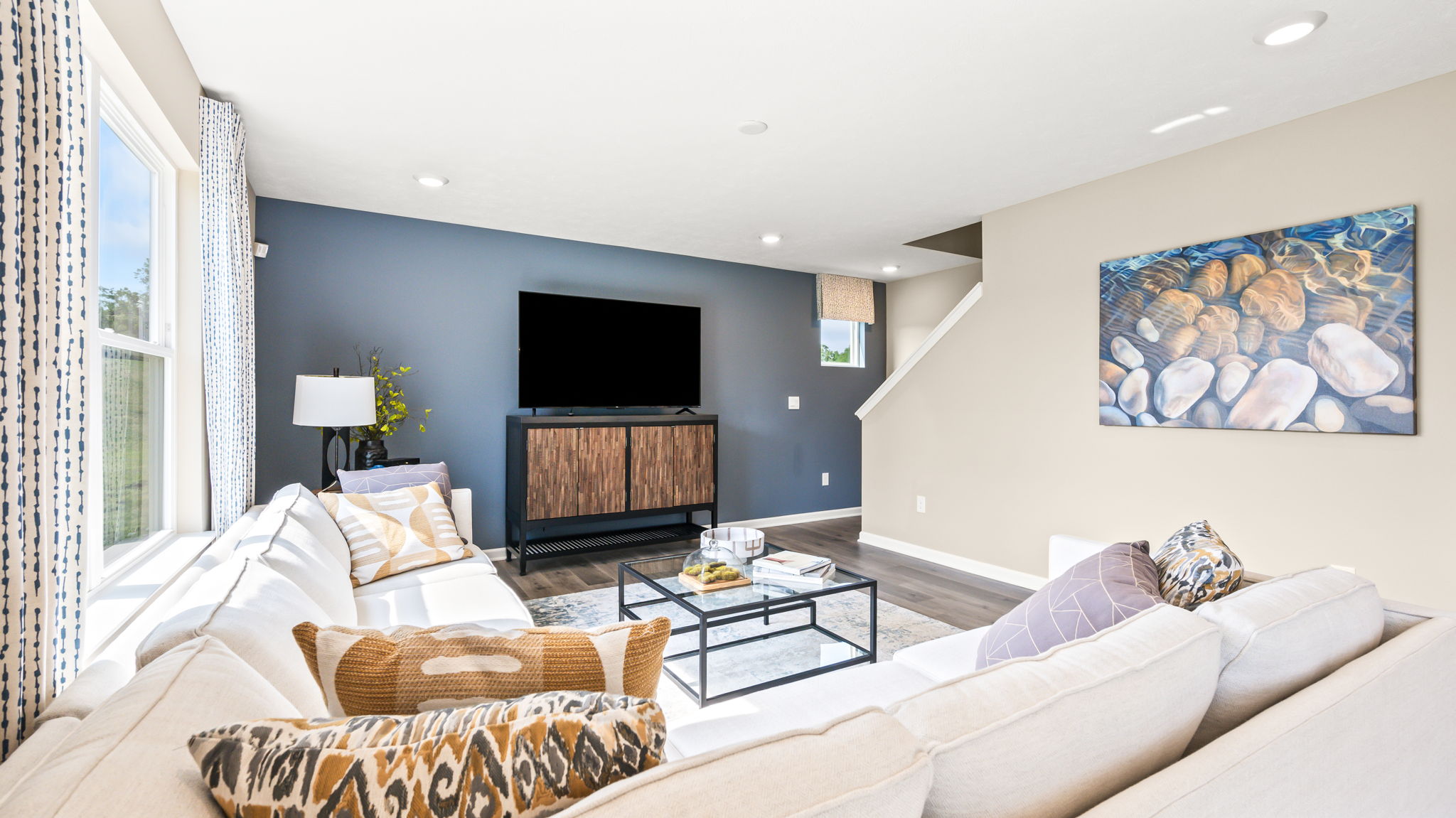 Living room with white sectional sofas, patterned pillows, glass coffee tables, a television, and artwork on neutral and blue walls.