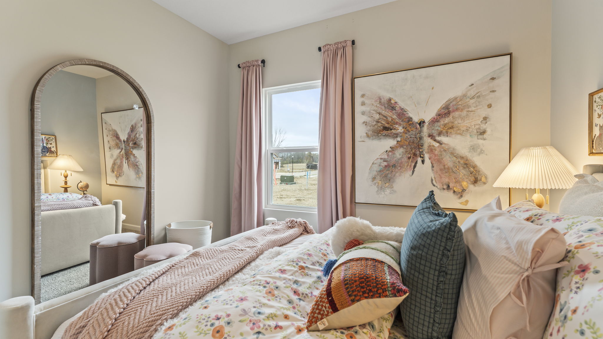 Side-view of bed with floral print, large butterfly art on wall, and rounded full-body mirror.
