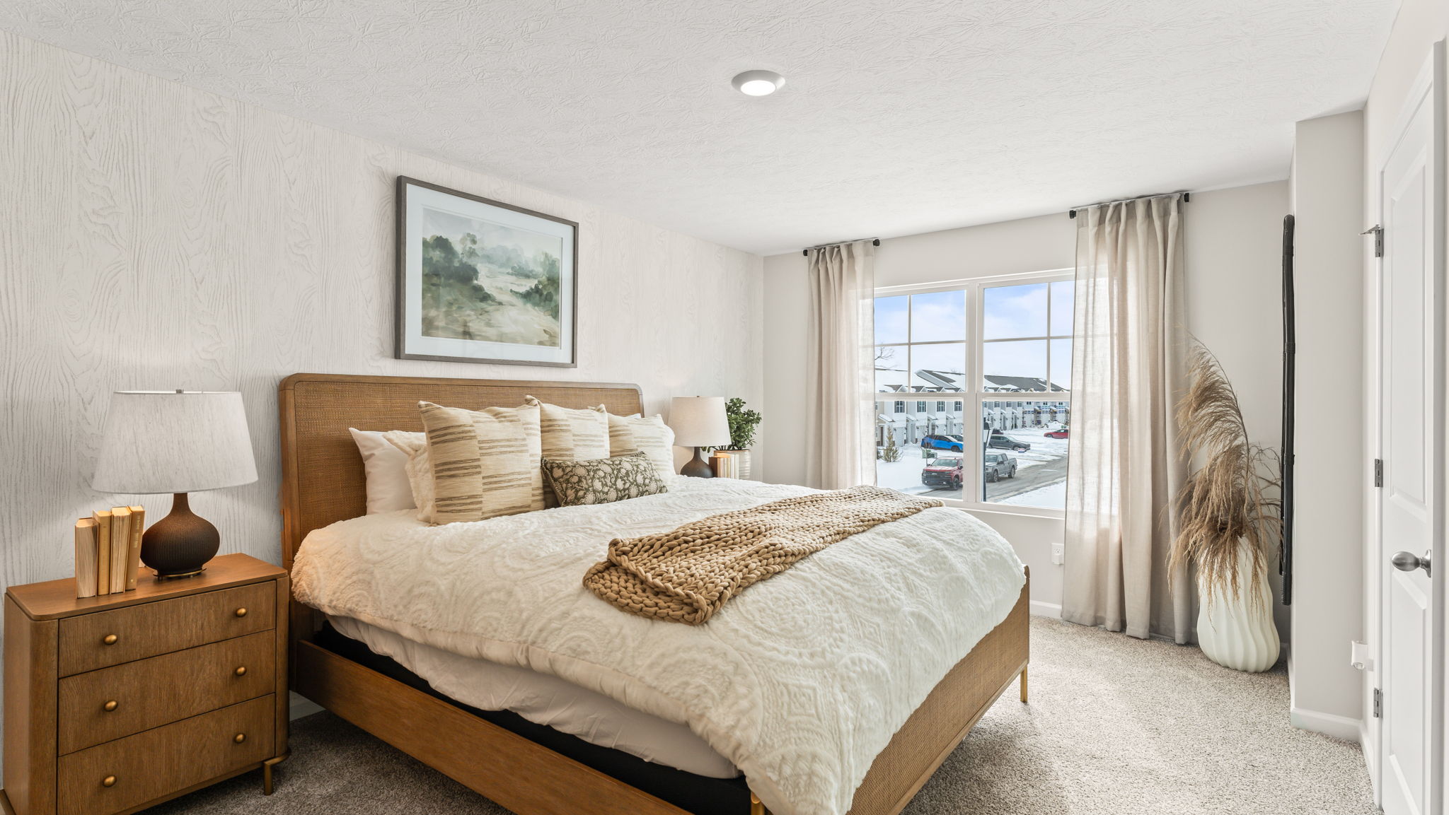 Primary bedroom with light bedding accented by oak wood colored furniture, plant, and drapery