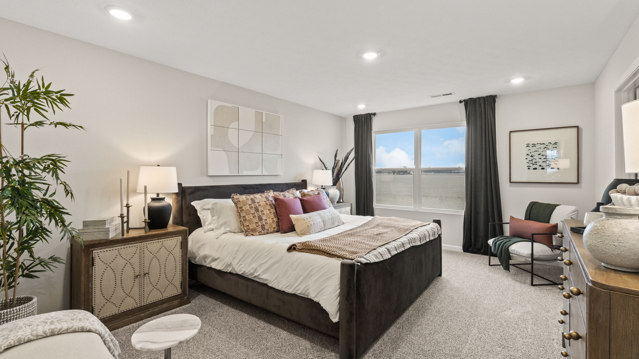 Full primary bedroom with neutral tones, plant, bed, chairs, and window with natural lighting.