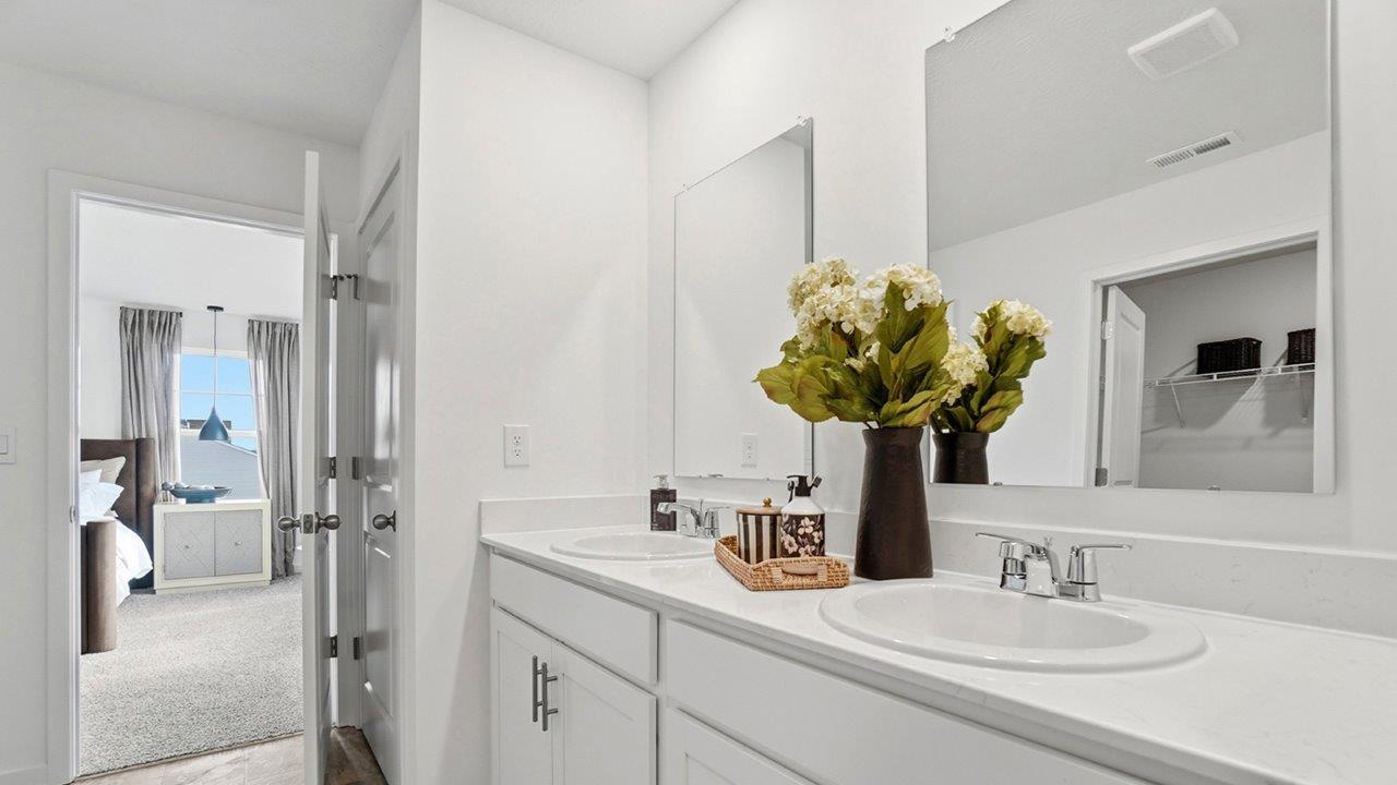 Bright double-sink bathroom vanity with white cabinetry, large mirrors, chrome faucets, and a vase of flowers.