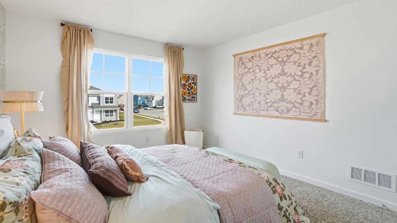 Bedroom view from the bed toward a large window, beige curtains, framed textile art, and neighboring homes outside.