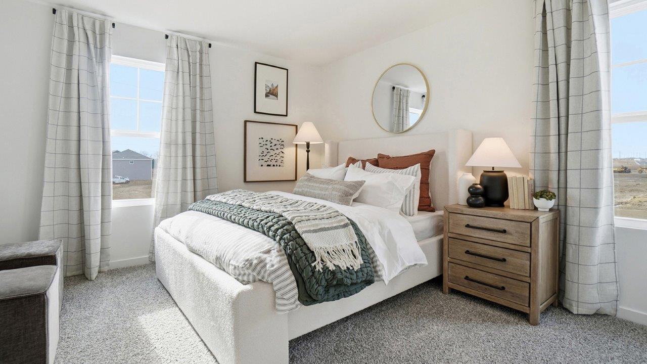 Light-filled bedroom with an upholstered white bed, green throw, wood nightstand, round mirror, and two windows.