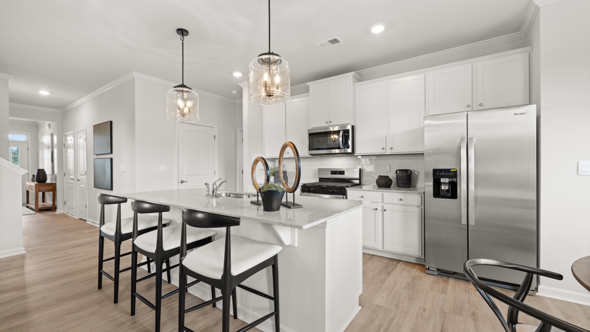 Three-quarter perspective of kitchen with pendant lights over island, stainless steel appliances, and chairs in front of the island.
