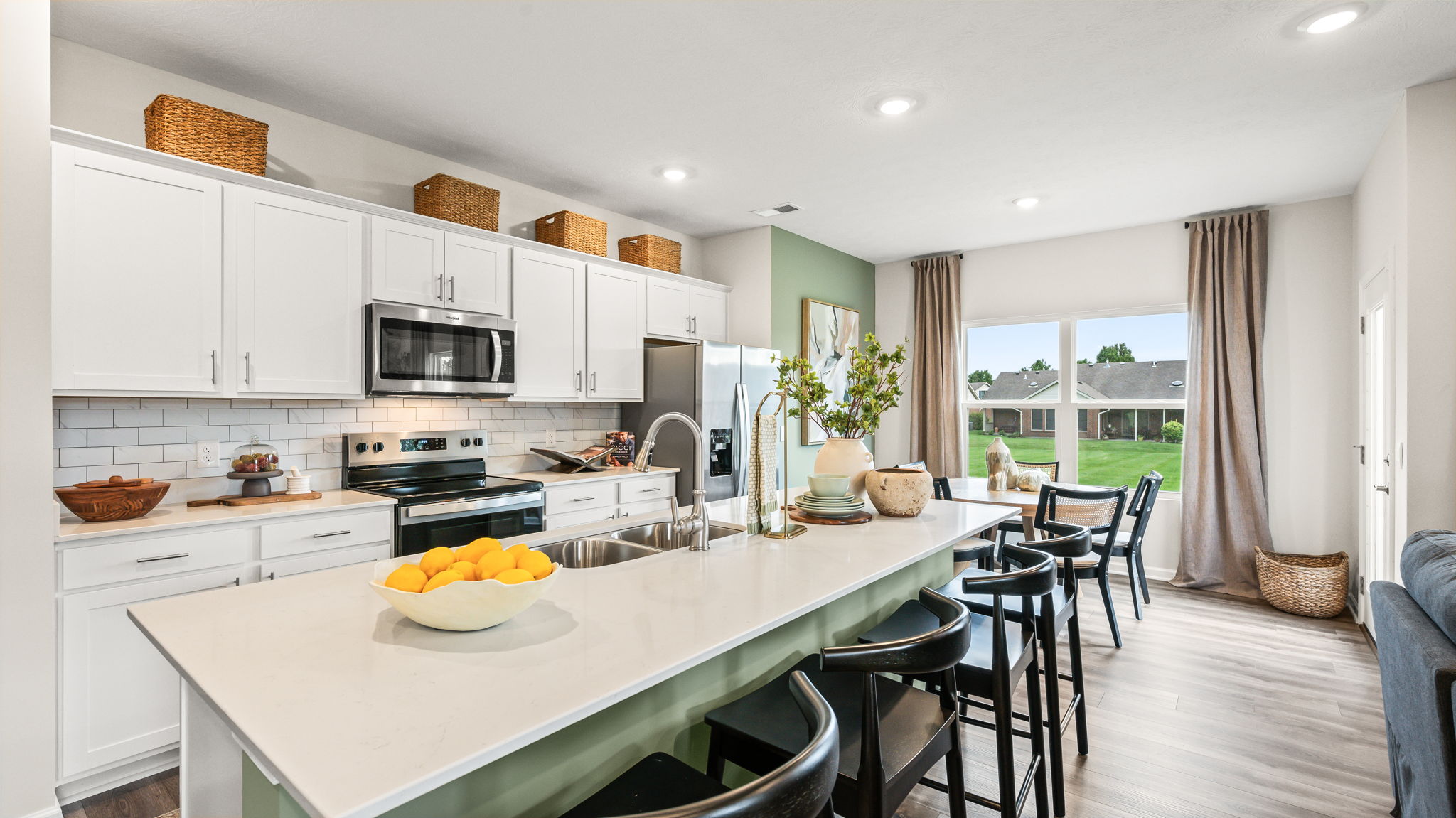 Kitchen with white shaker cabinets, stainless steel appliances, woven baskets above the cabinets, a long pale green island with black bar stools, a bowl of oranges, and a dining area by tall curtained windows.