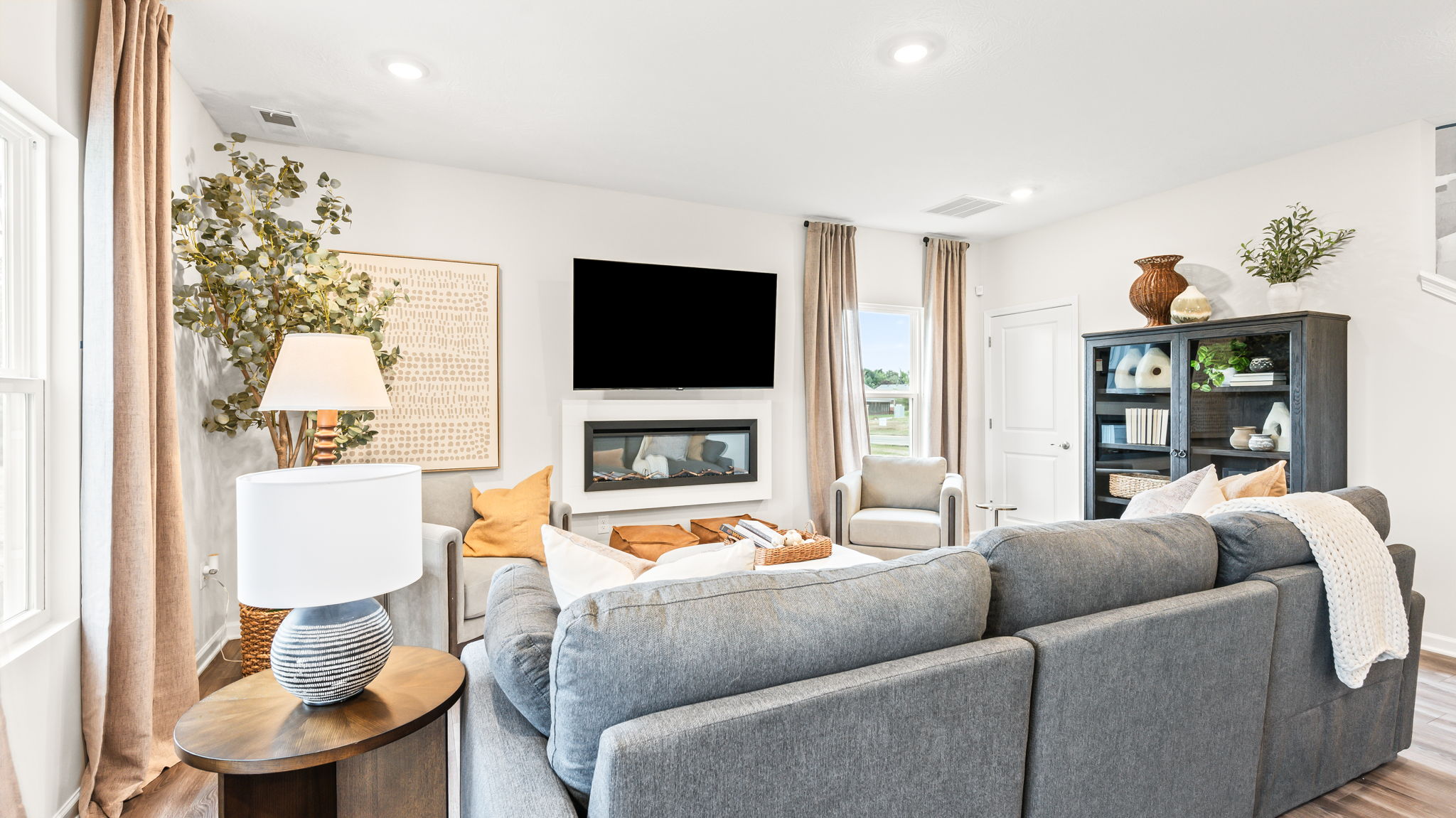Living room viewed from behind a gray sectional, with a wall-mounted television above a linear fireplace, two light armchairs, tall curtained windows, a floor lamp, artwork, and a dark display cabinet.