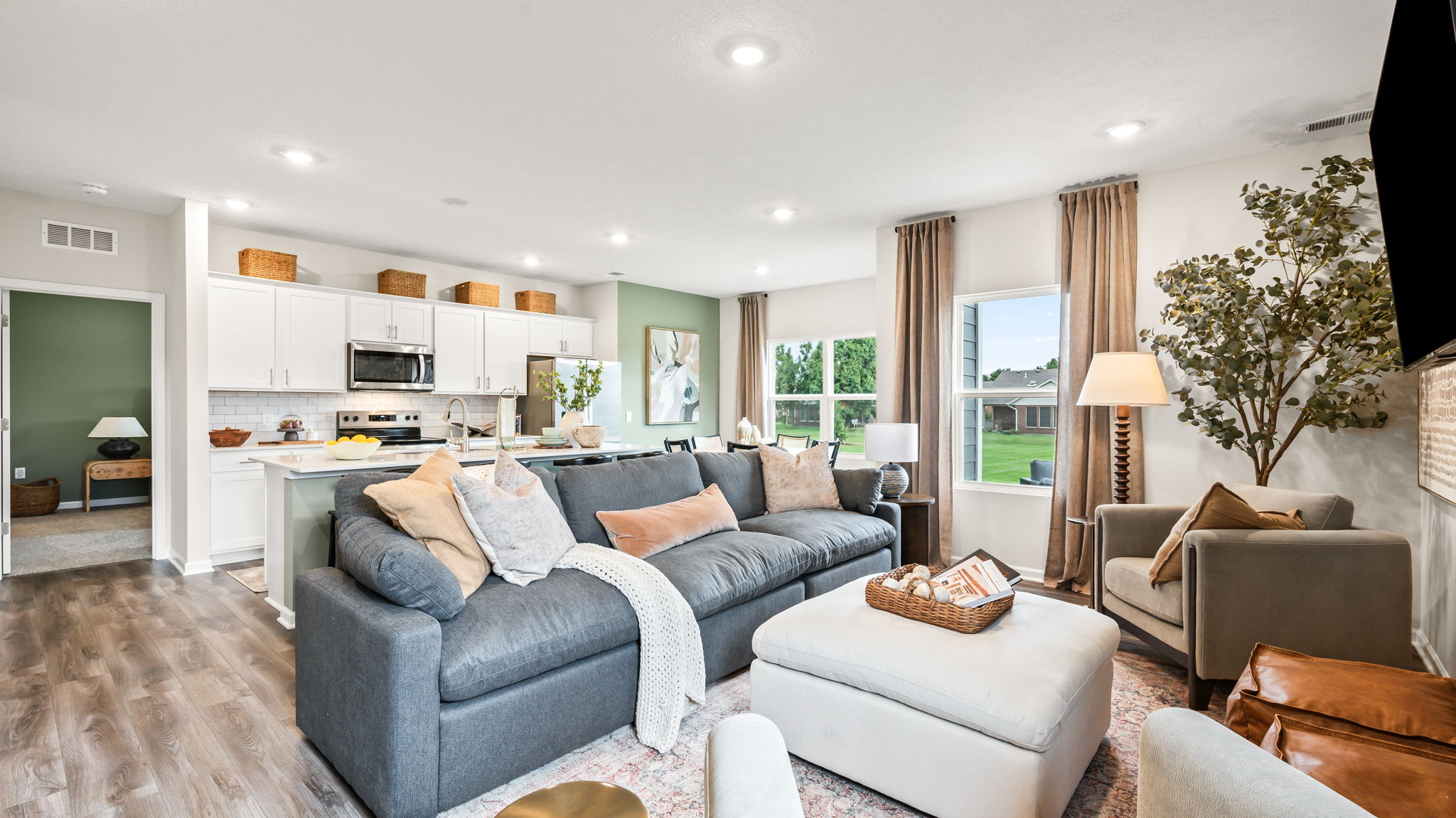 Open-plan living room and kitchen with a gray sectional, white ottoman, armchairs, tall curtained windows, and a white kitchen with an island in the background.