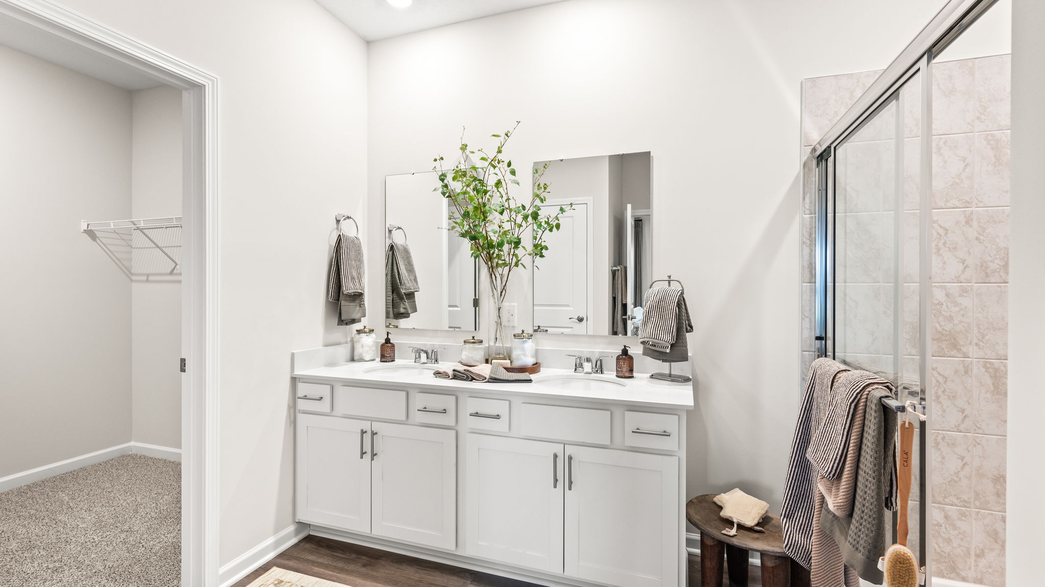 Bathroom with a white double-sink vanity, large mirrors, chrome faucets, striped towels, decorative branches, an adjoining carpeted walk-in closet, and a glass-enclosed beige tile shower.