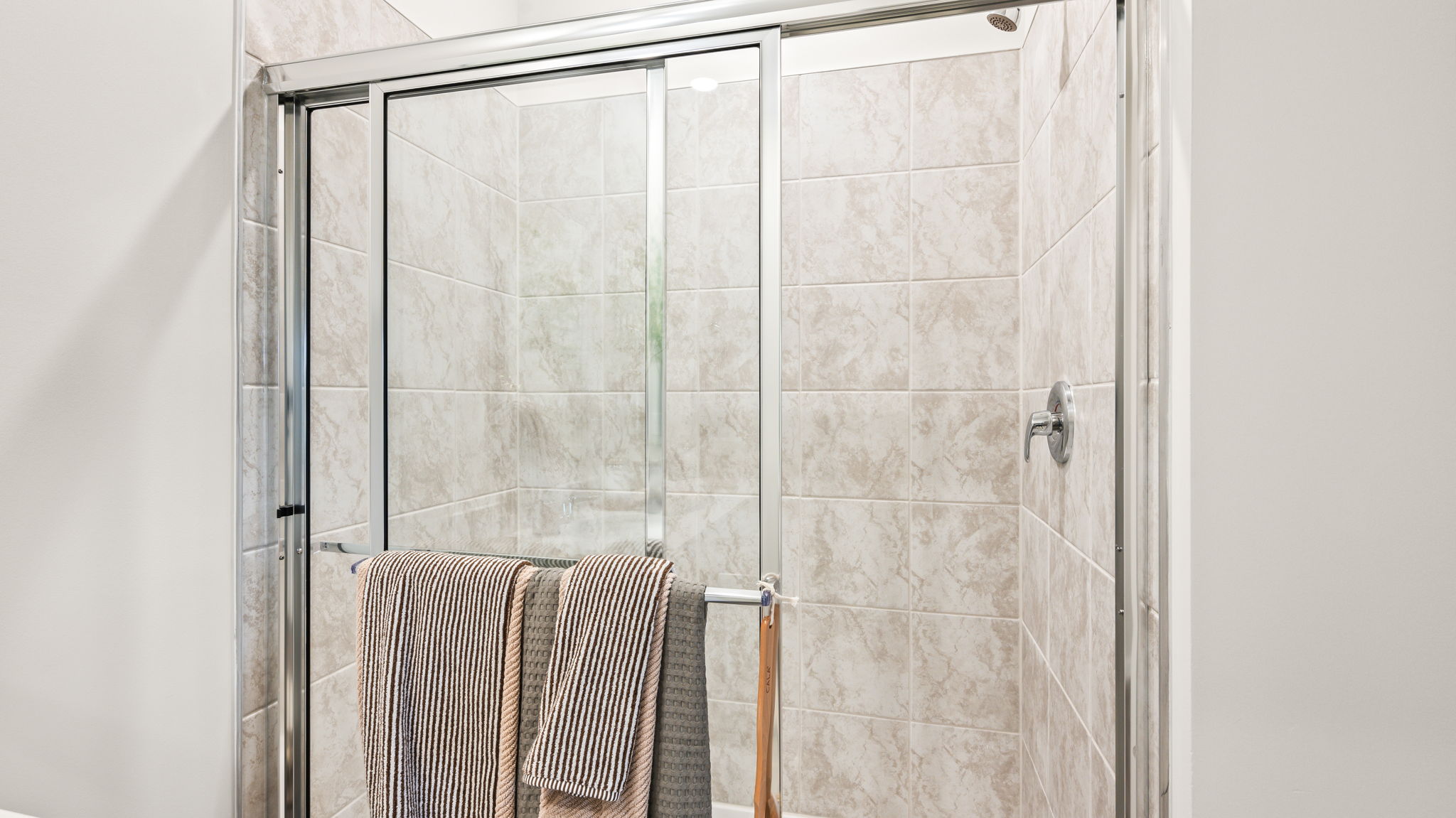 Close view of a glass-enclosed shower with beige tiled walls, chrome fixtures, and striped towels hanging across the sliding shower door.