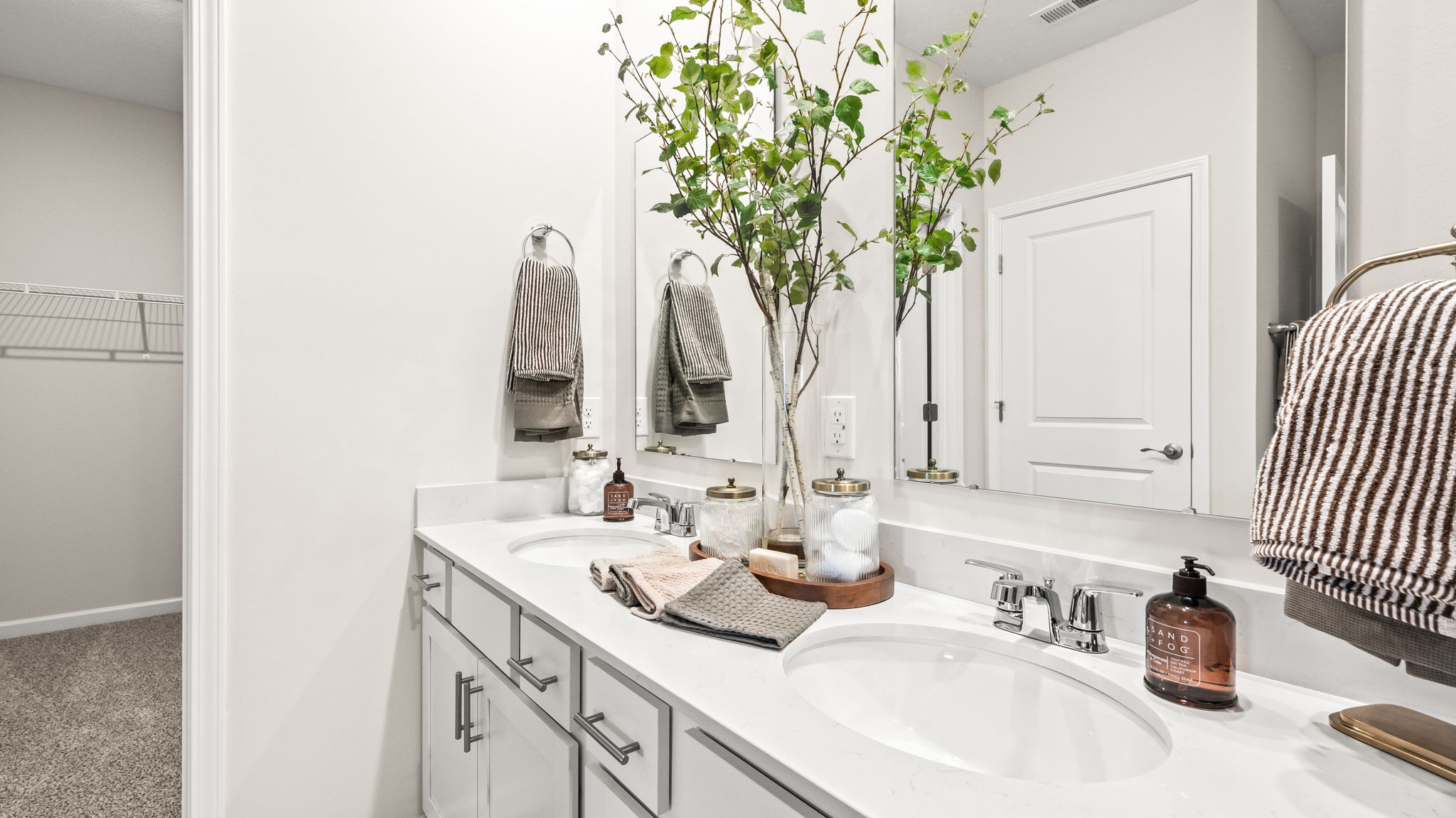 Close view of a bathroom double-sink vanity with white cabinetry, chrome faucets, large mirrors, striped towels, folded washcloths, glass jars, and tall decorative branches beside an open closet.