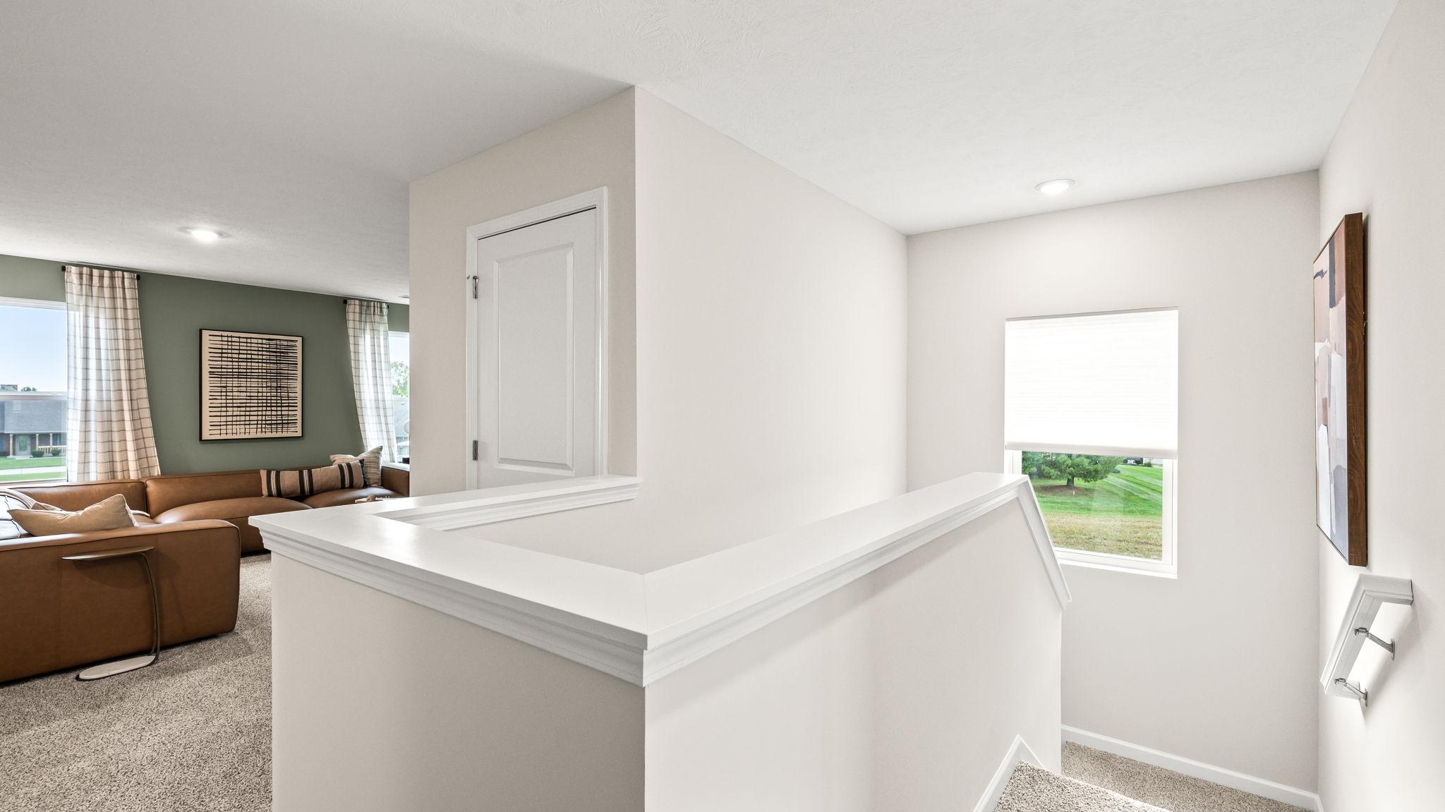 Upstairs landing with white railings around a stairwell, a window with a white shade, carpeted flooring, and an adjacent sitting area with a tan sectional and green accent wall.