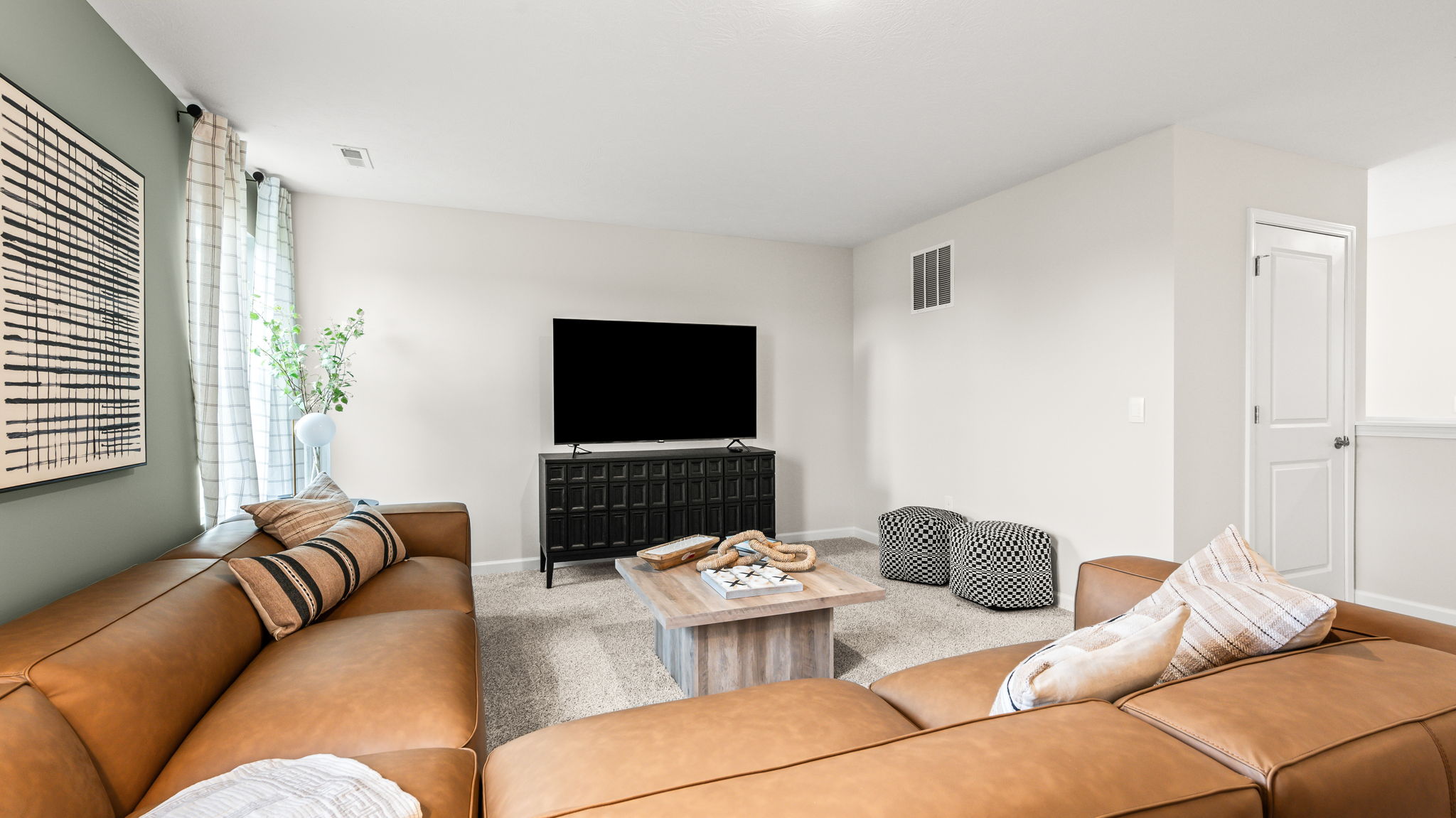 Bonus room seating area with a tan sectional around a wood coffee table, television on a dark media console, two patterned ottomans, tall windows, and carpeted flooring.
