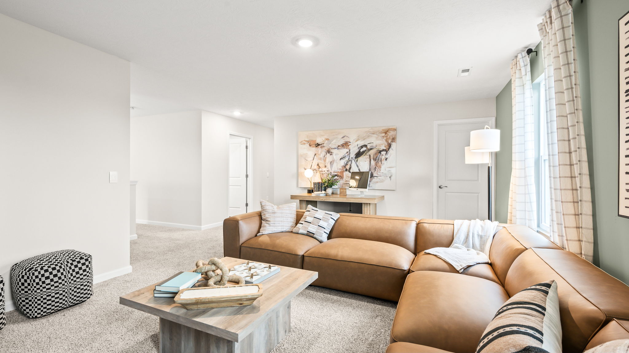 Bonus room viewed across a tan sectional and wood coffee table toward a desk beneath abstract artwork, with tall curtained windows and neutral carpet.
