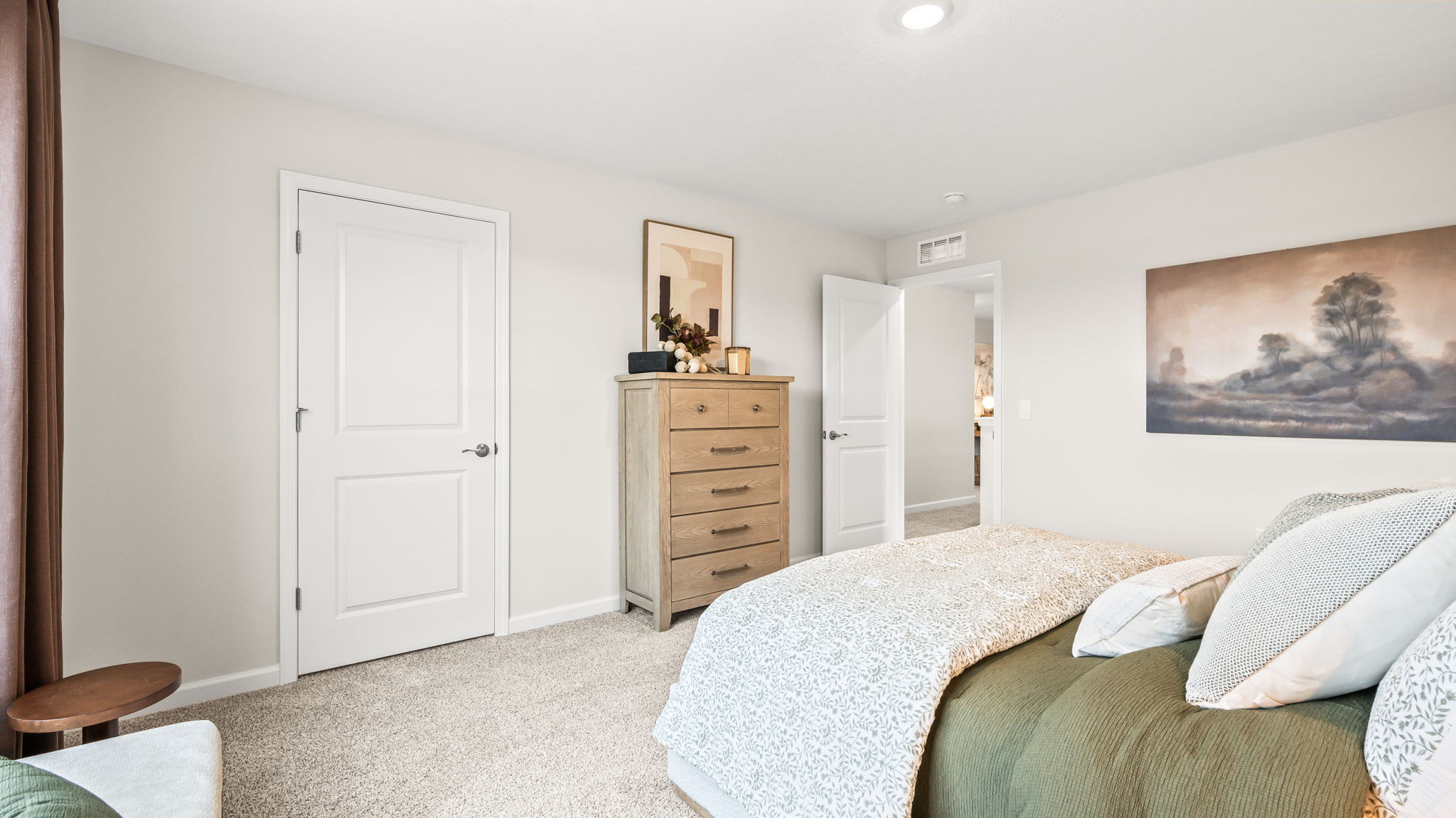 Bedroom viewed toward a tall wood dresser, two white doors, landscape artwork, and the side of a bed with green and cream bedding on neutral carpet.