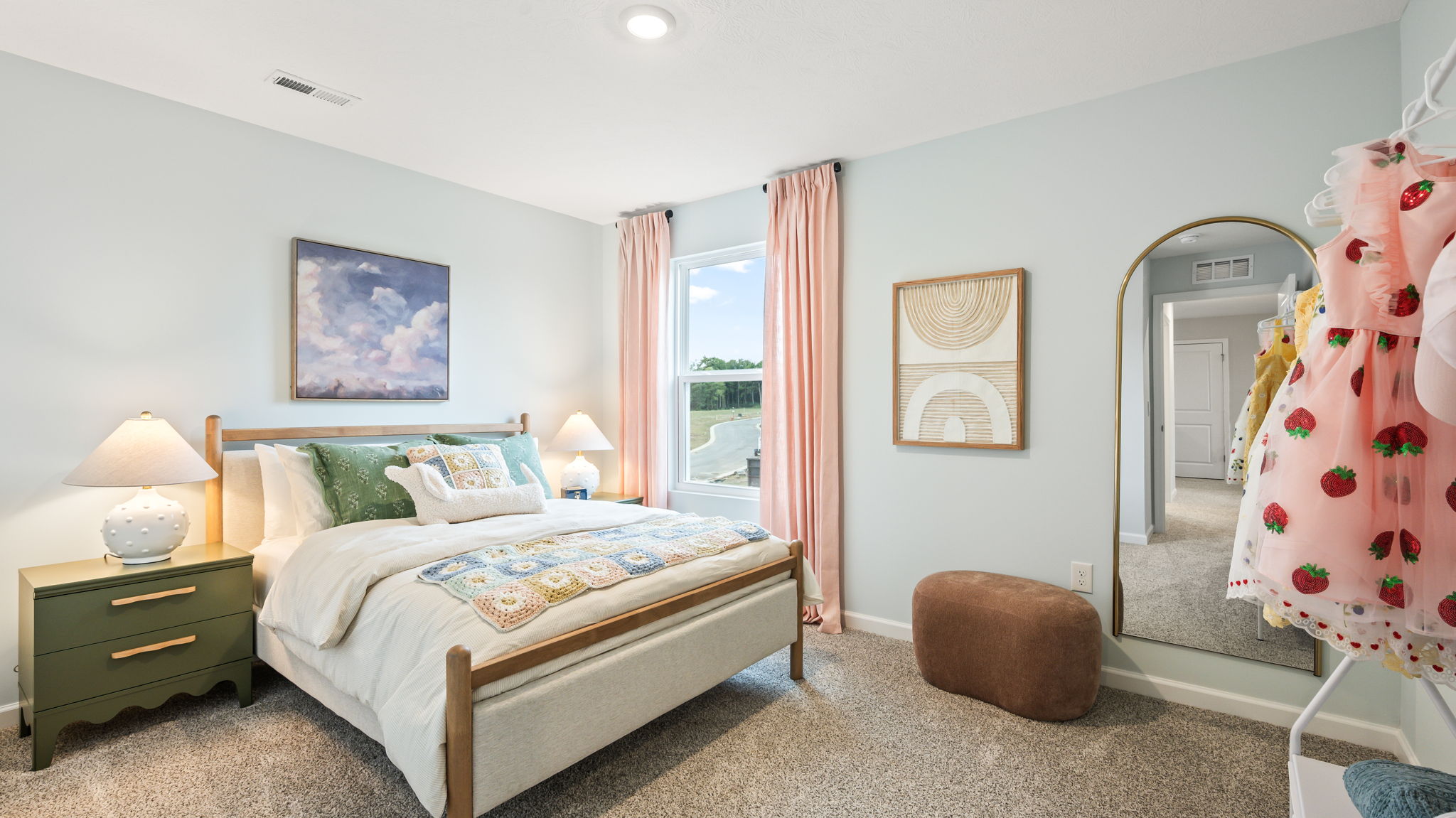 Bedroom with pale blue walls, a wood-framed bed with colorful patterned bedding, green nightstands, pink curtains, a tall arched mirror, framed art, and a clothing rack.