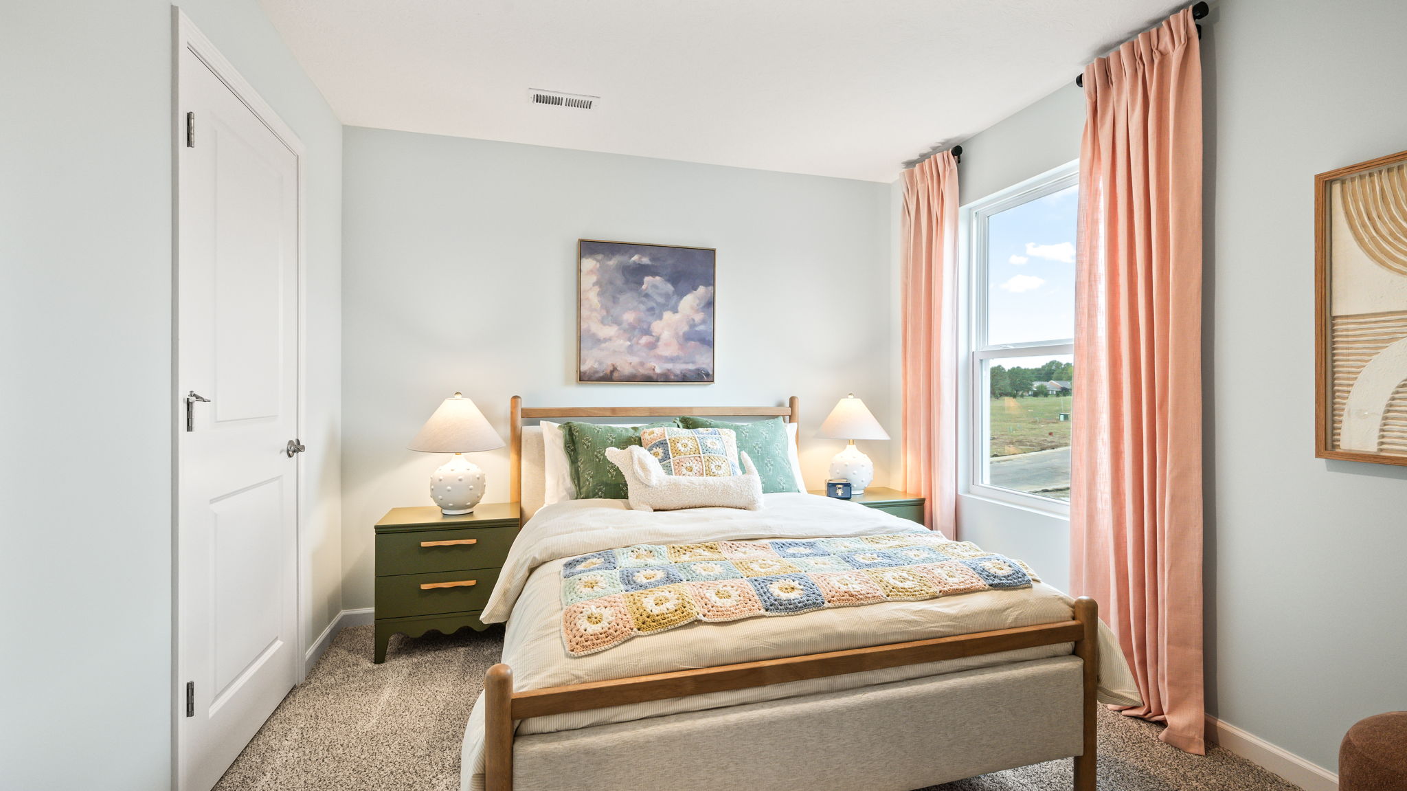 Front view of a bedroom with a wood-framed bed, colorful patterned bedding, green nightstands and lamps, pale blue walls, framed art, and pink curtains around a window.