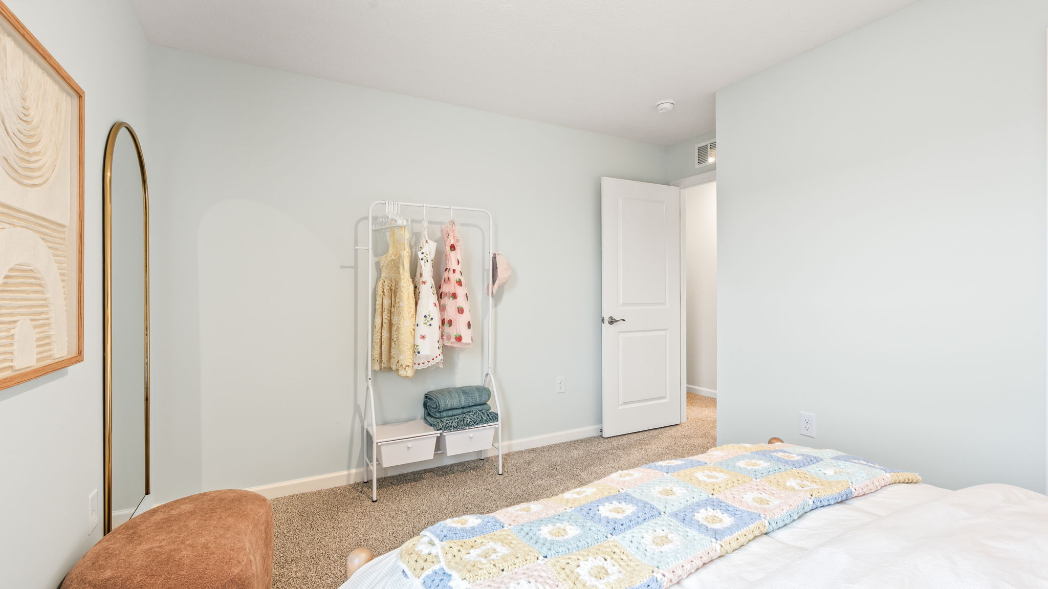Bedroom corner with pale blue walls, a tall arched mirror, white clothing rack holding children's dresses, a small bench with folded linens, and the foot of a patterned bed.