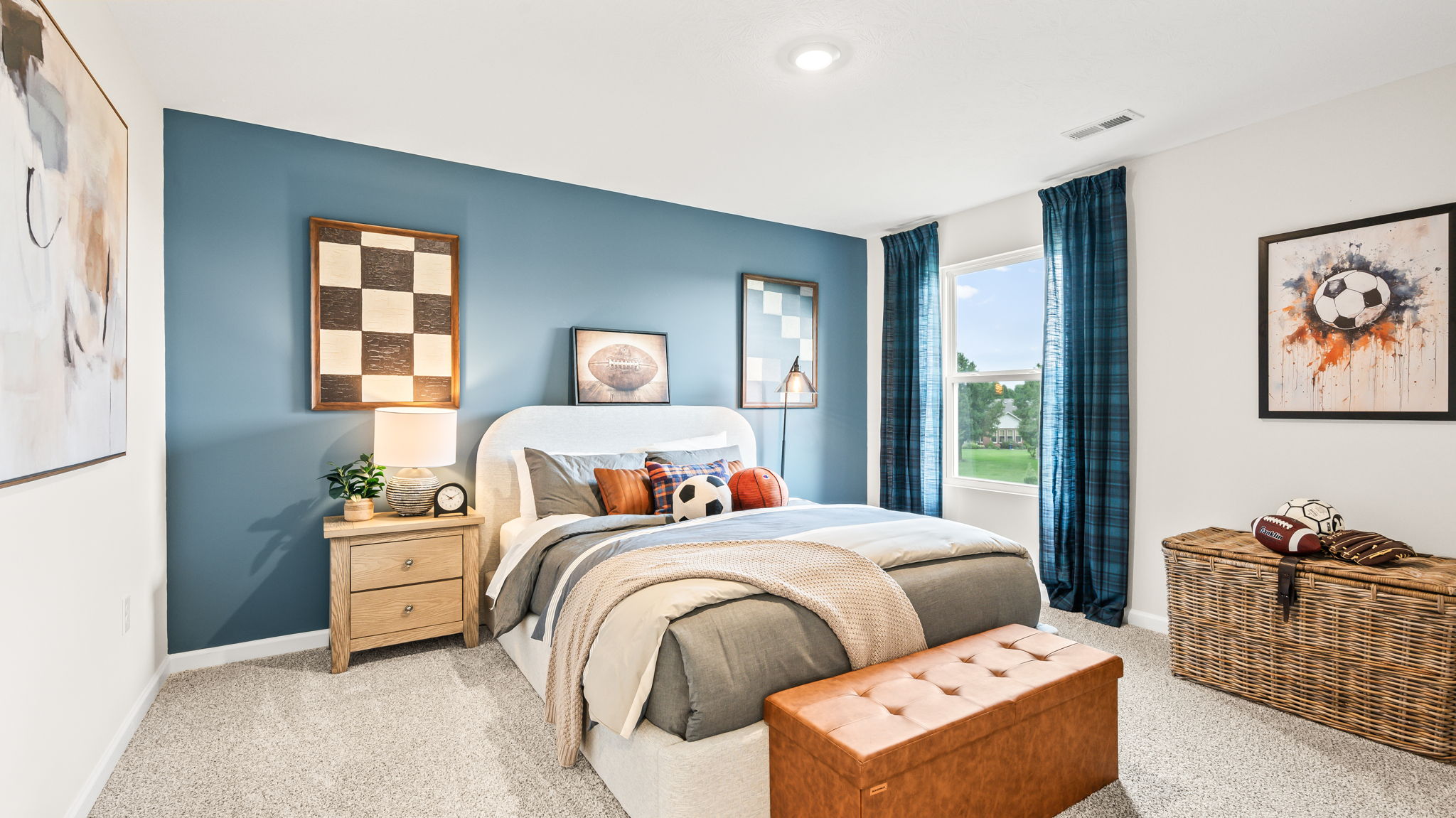 Bedroom with a blue accent wall, upholstered bed, gray and blue bedding, sports-themed pillows and artwork, dark teal curtains, wood nightstand, leather bench, and woven storage chest.