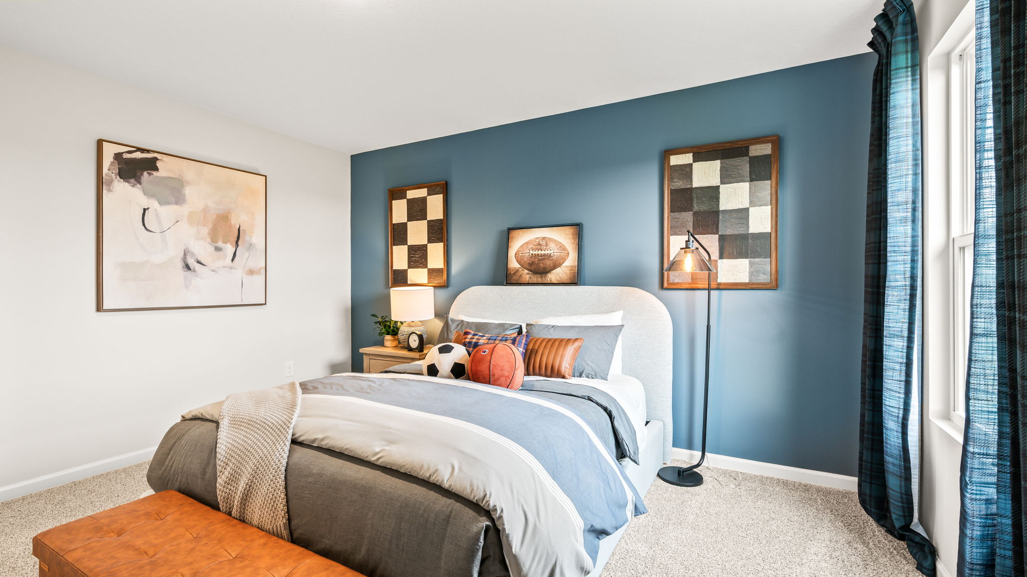 Bedroom viewed toward a blue accent wall with an upholstered bed, gray and blue bedding, sports-themed pillows and artwork, a floor lamp, leather bench, and dark teal curtains.