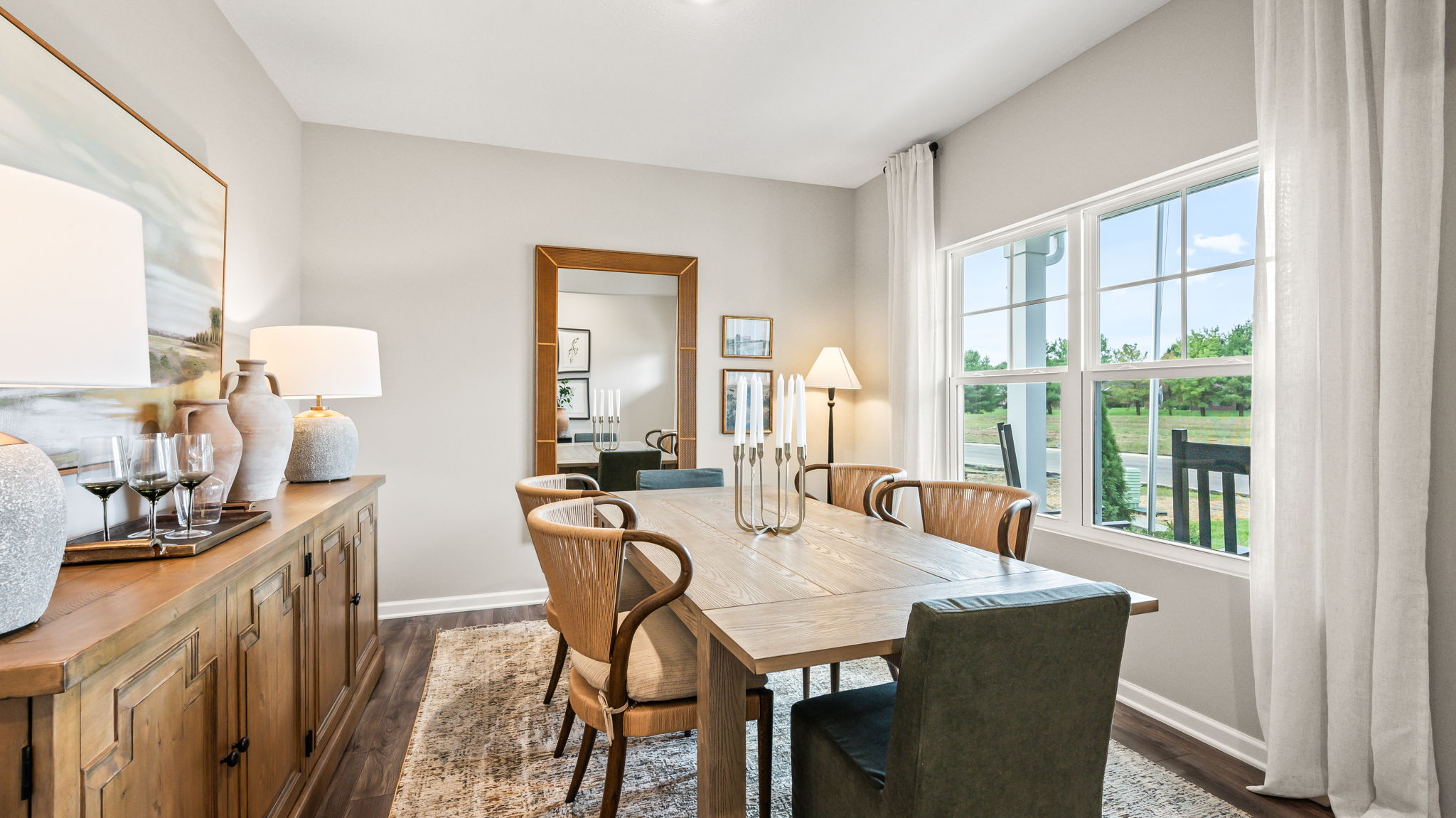 Dining room with a rectangular wood table, mixed upholstered and woven chairs, a large wall mirror, wood sideboard with lamps and decor, tall white curtains, and a large window.