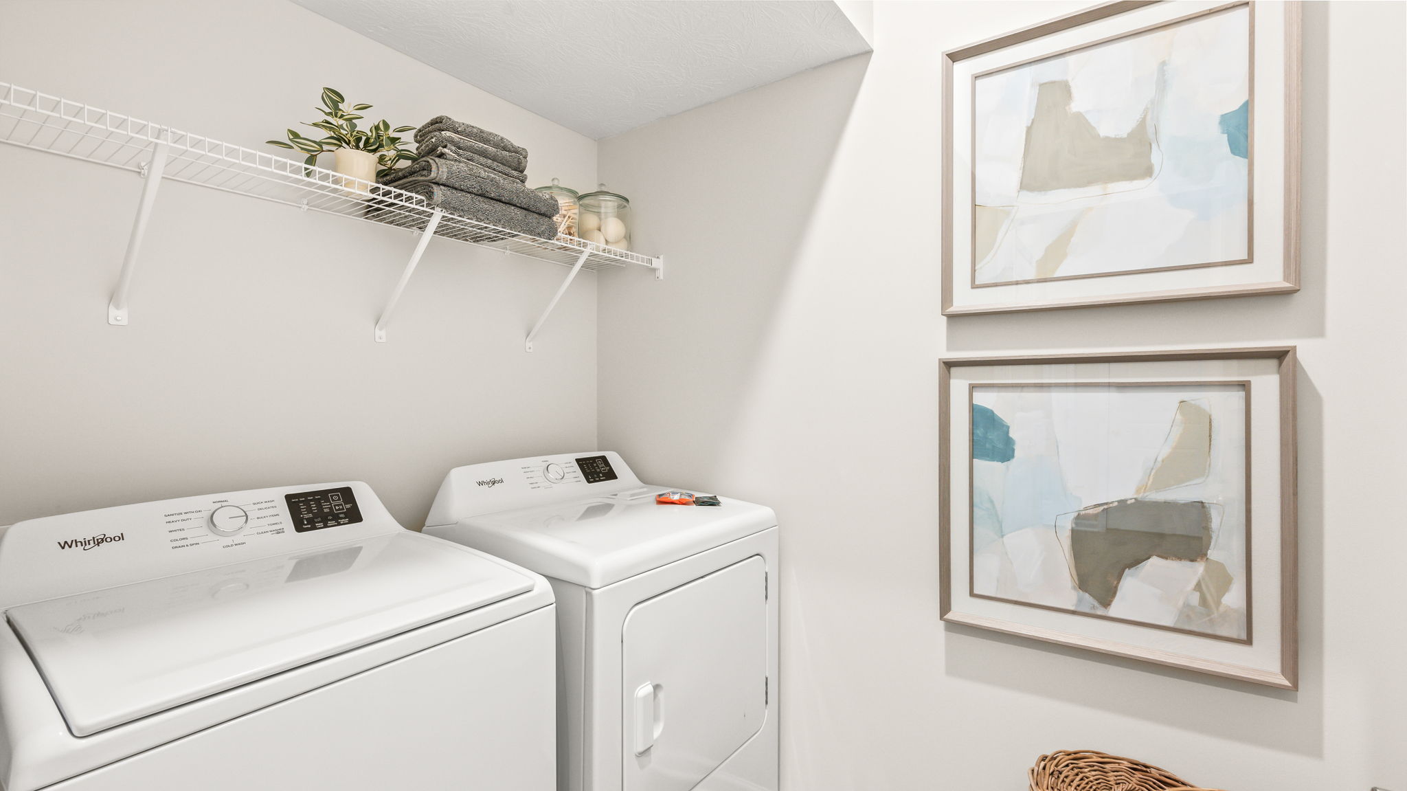 Laundry room with a white top-loading washer and front-loading dryer, white wire shelf holding folded towels and containers, two framed abstract prints, and a woven laundry basket.
