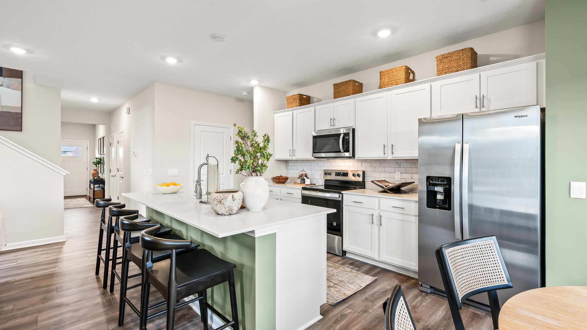 Kitchen viewed from the dining area, with white shaker cabinets, stainless steel appliances, a pale green island with black bar stools, woven baskets, and wood-look flooring.