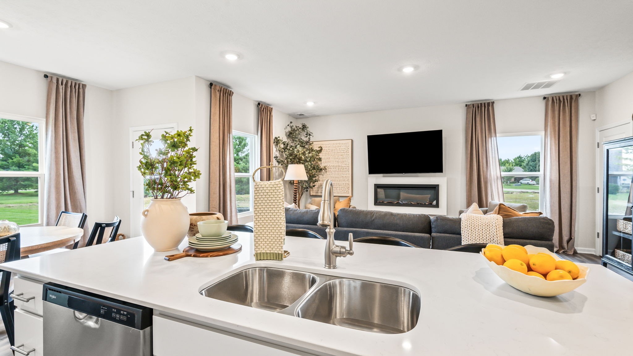 View from a white kitchen island with a double sink and bowl of oranges toward a dining area and living room with a gray sectional, wall-mounted television, fireplace, and tall windows.