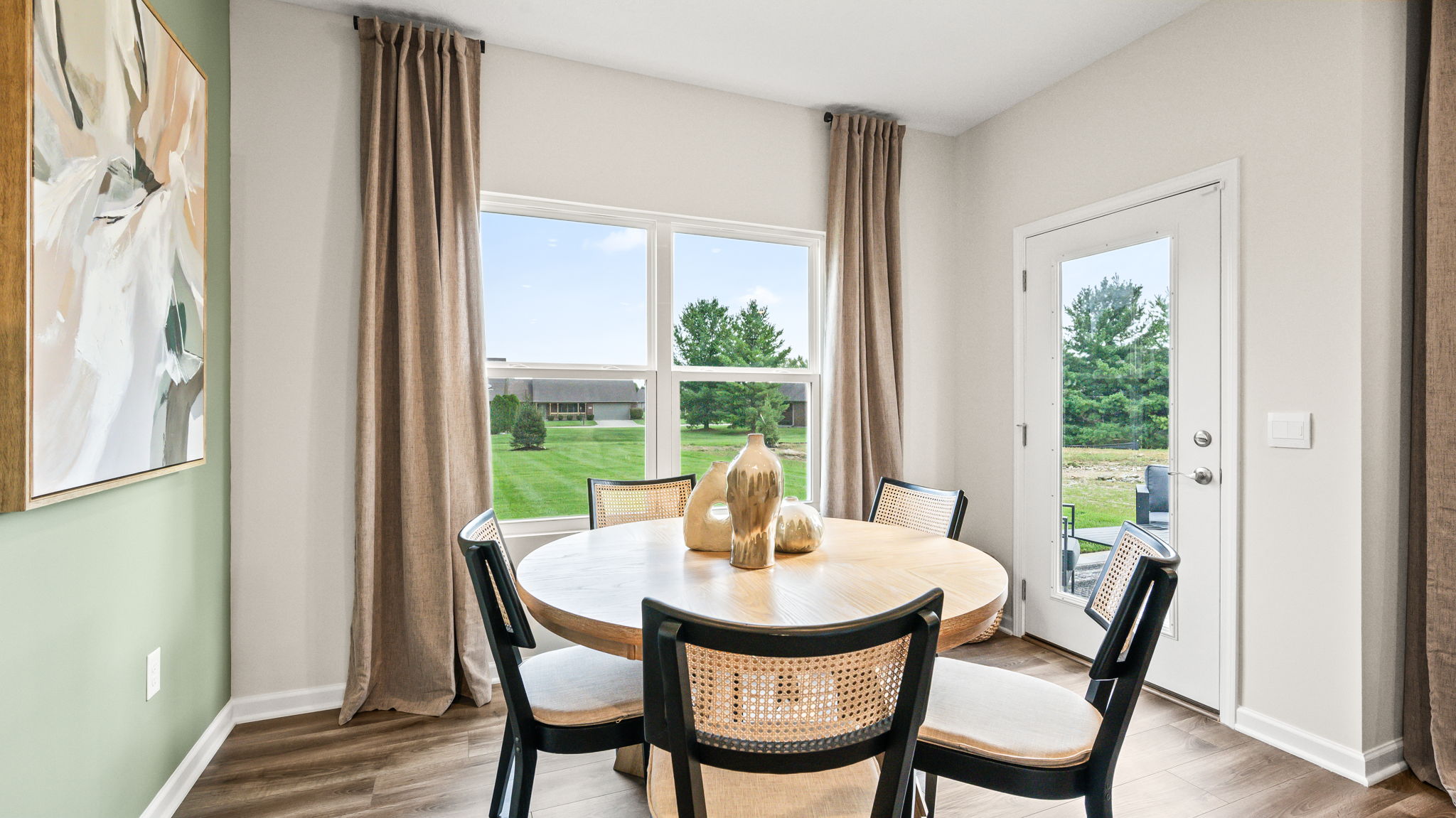 Dining nook with a round wood table, six black cane-back chairs, decorative vases, tall curtained windows, a green accent wall with abstract artwork, and a glass-paneled exterior door.