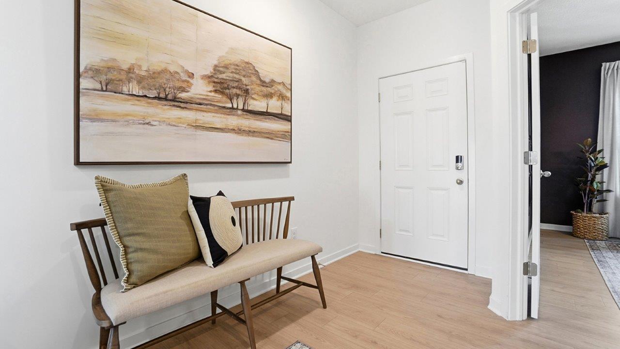 Entryway with a wood bench, neutral pillows, landscape artwork, light wood flooring, and adjacent doorway.