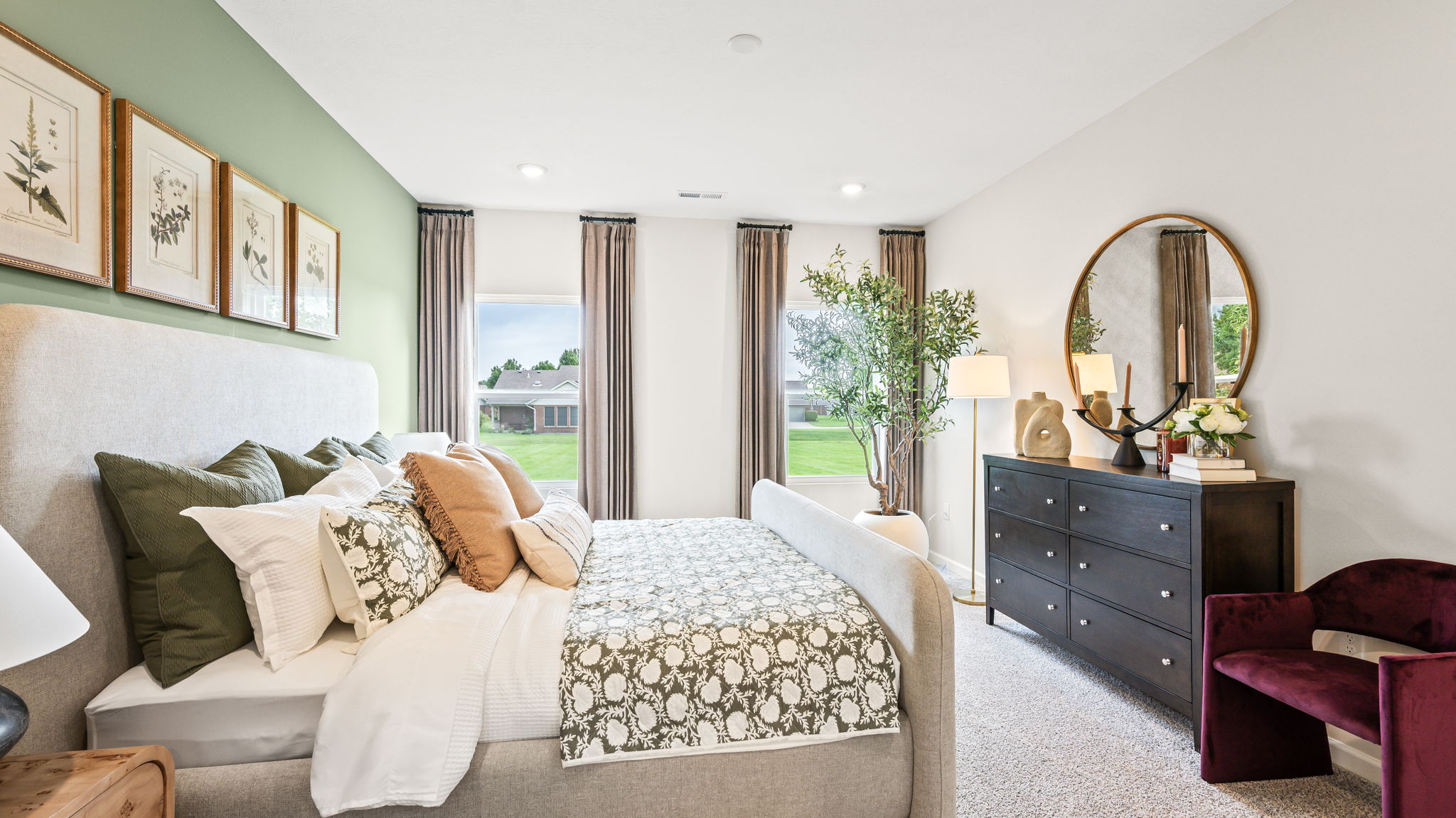 Bedroom viewed from beside an upholstered bed, with green patterned bedding, layered pillows, botanical prints on a green accent wall, a dark dresser, round mirror, decorative tree, and burgundy chair.