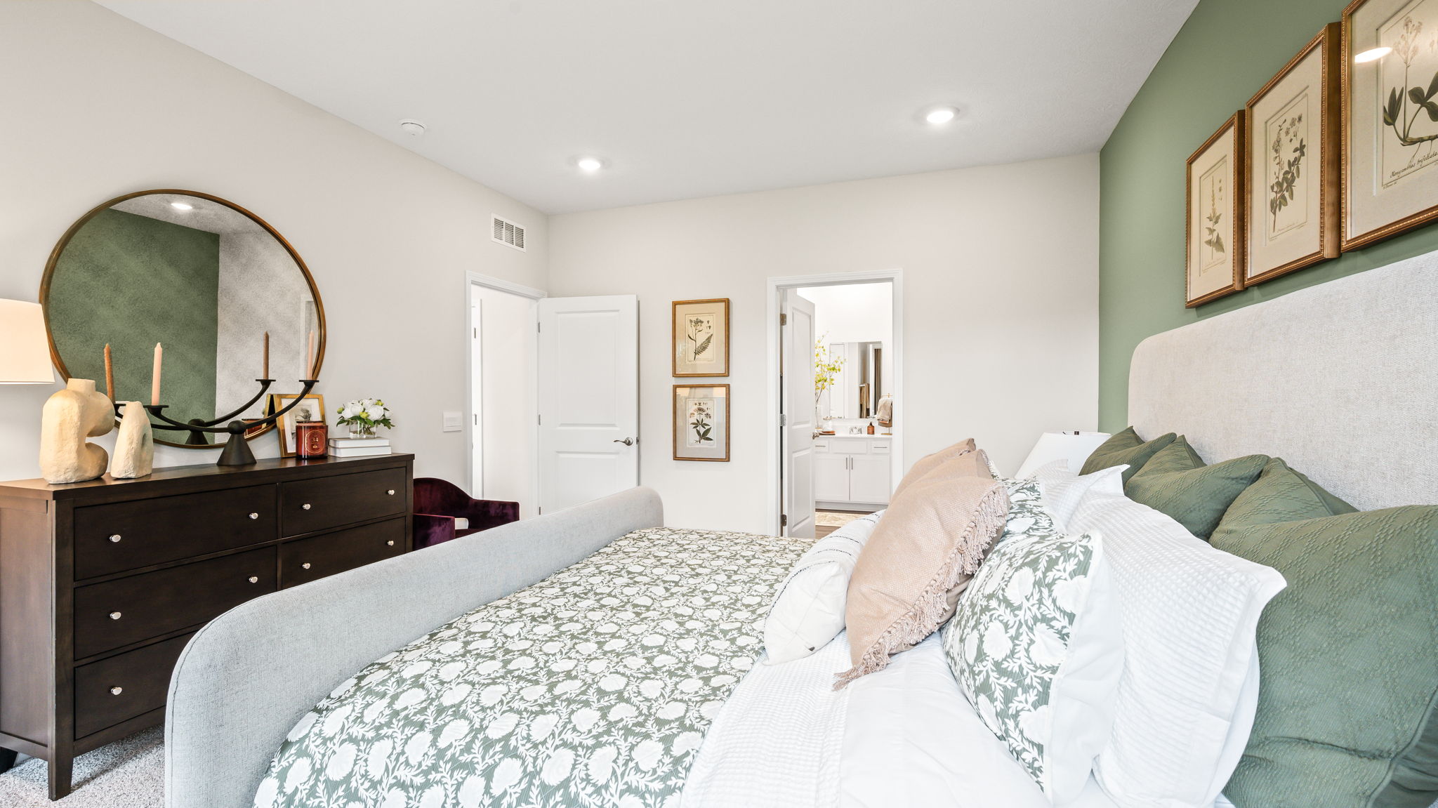 Bedroom viewed from the foot and side of an upholstered bed, with green patterned bedding, layered pillows, a dark dresser, round mirror, botanical artwork, and an open doorway to the bathroom.