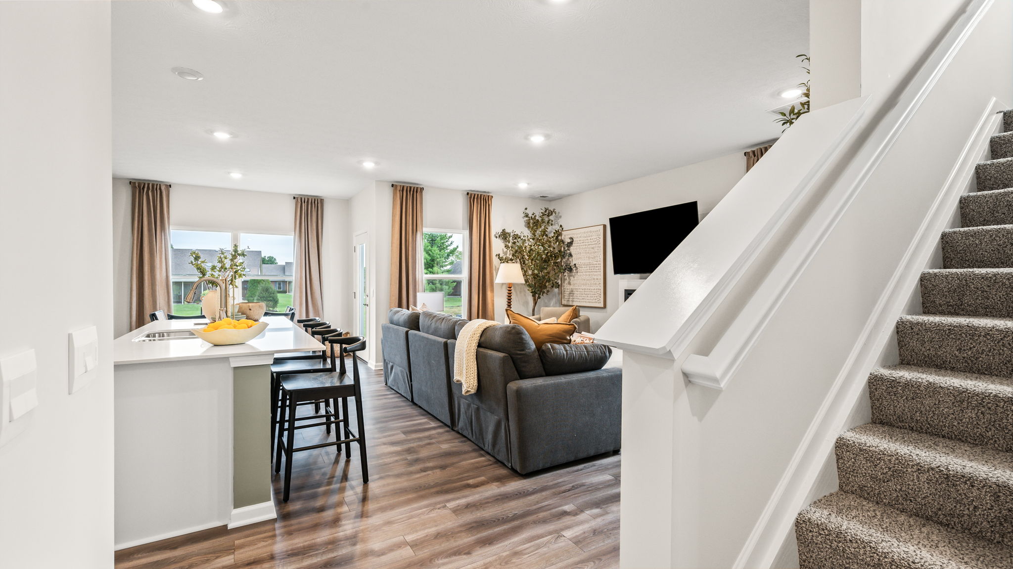 Open-plan kitchen and living area viewed beside a carpeted staircase, with a white island, black bar stools, gray sectional, wall-mounted television, fireplace, and wood-look flooring.