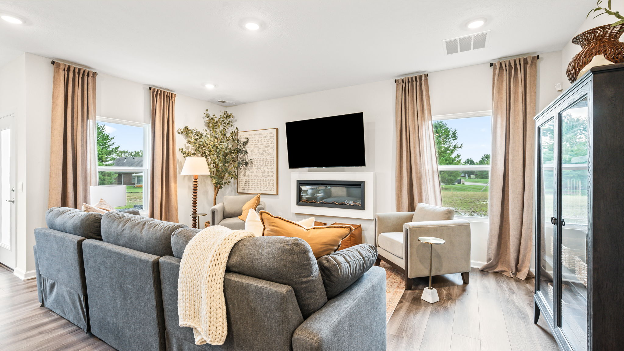 Living room with a gray sectional facing a wall-mounted television and linear fireplace, two light armchairs, tall curtained windows, a floor lamp, artwork, and a dark glass-front display cabinet.